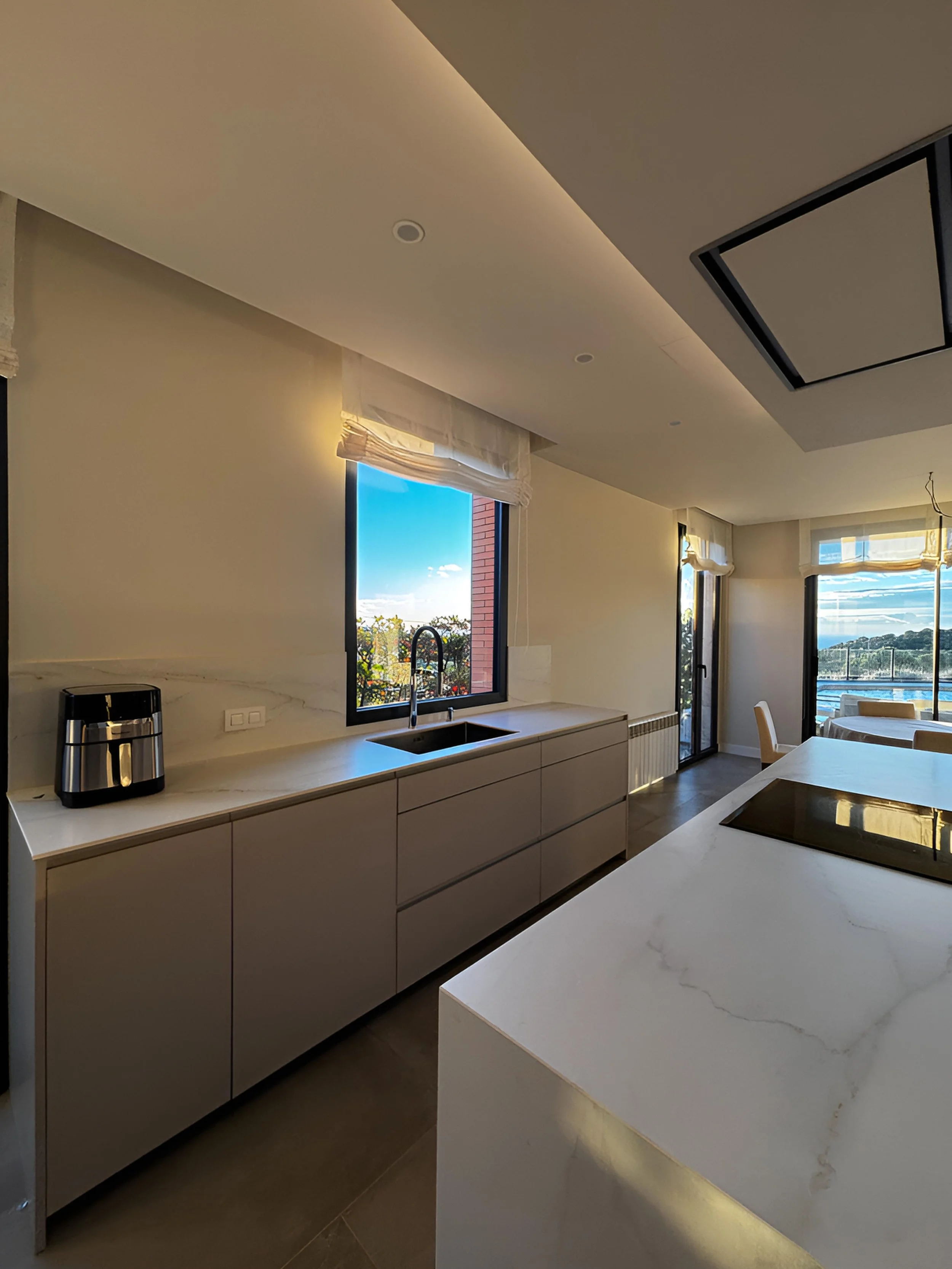 Cocina moderna con encimera de piedra, fregadero, ventanas grandes con vista al exterior, y electrodomésticos.