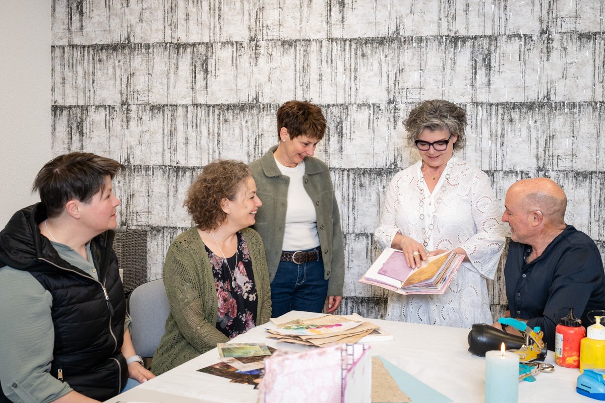 Eine Gruppe von fünf Menschen, drei Frauen und zwei Männer, sitzen und stehen um einen Tisch mit Papieren und Farben. 	
Petra Natalie Fahrni in weisser Kleidung zeigt auf ein Buch, während die anderen zuschauen und lächeln.