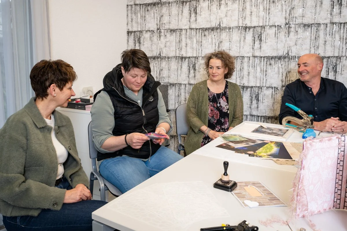 Vier Menschen sitzen an einem Tisch mit Fotos und Bastelmaterial. Sie sind an einem Bastel- oder Kreativprojekt beteiligt.
