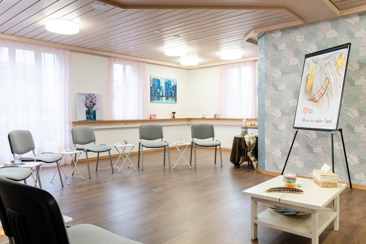 Ein heller, räumlicher Seminarraum mit Stühlen, die im Kreis um kleine Tische angeordnet sind. Der Raum ist mit Vorhängen, Gemälden und Kerzen dekoriert, es gibt eine Flipchart am rechten Rand.