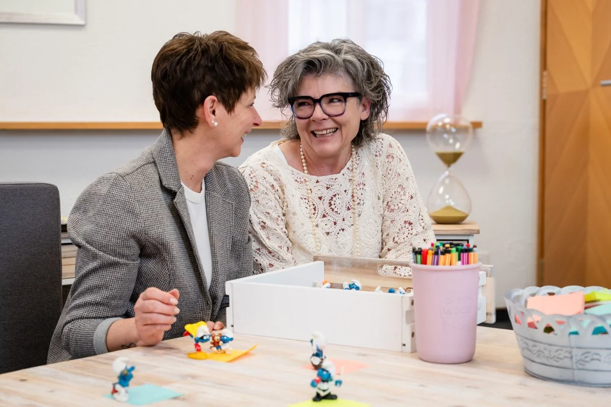 Zwei Frauen lachen und sprechen in einem Raum, umgeben von Bastelmaterialien wie Filzstiften und kleinen Figuren, in einer freundlichen Umgebung.