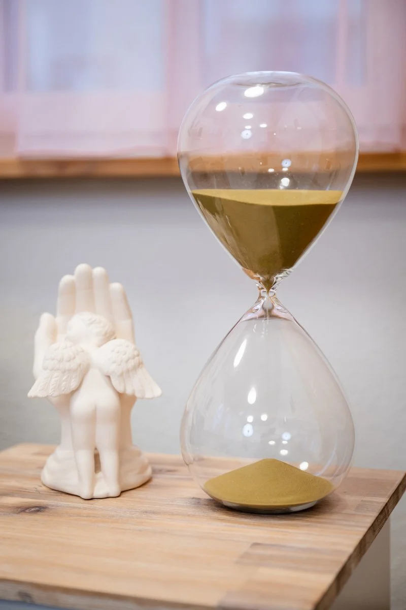 Sanduhr auf Holztablett neben einem Engel. Hintergrund mit durchscheinendem Vorhang.