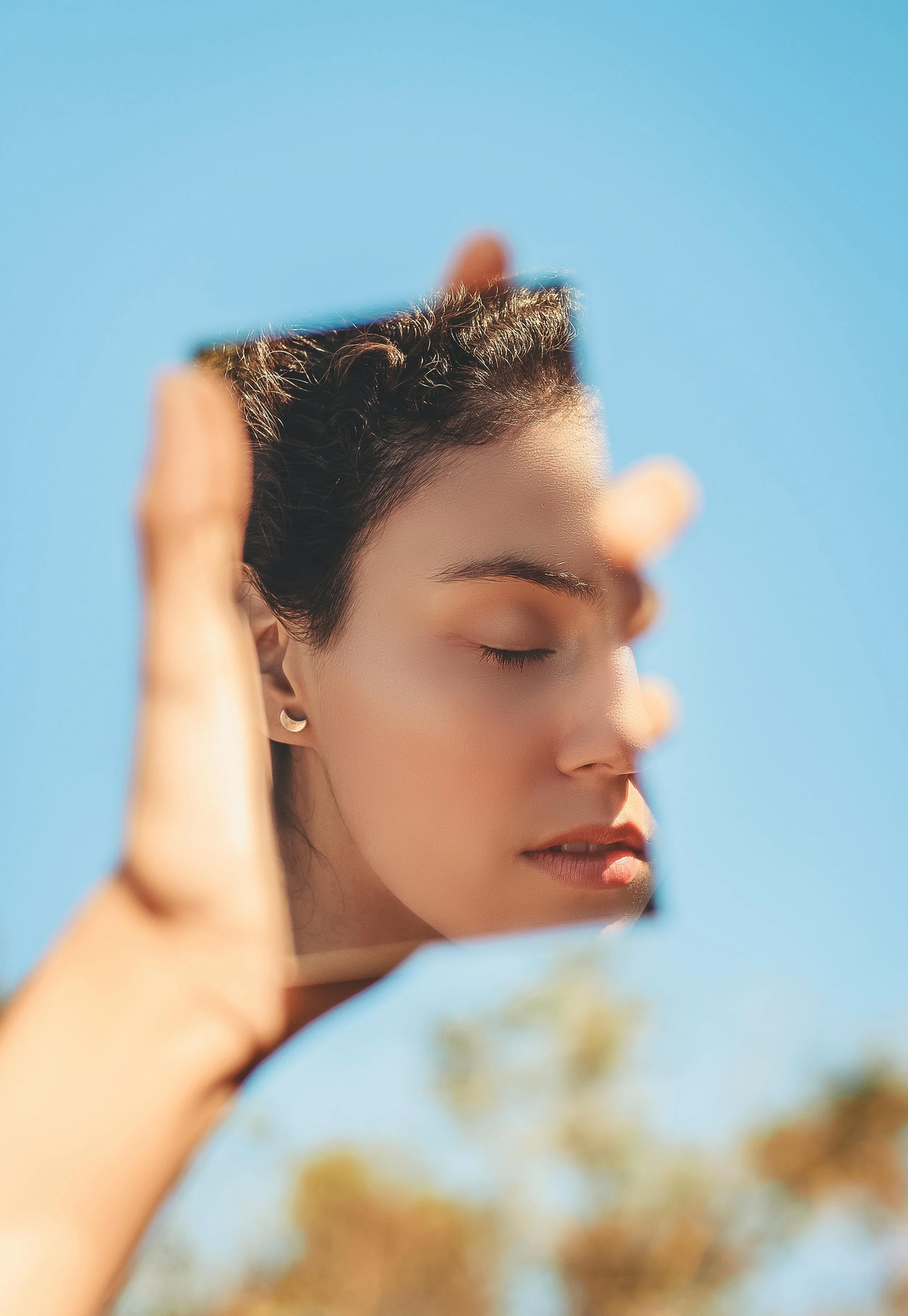 A young woman with dark, natural hair and small hoop earrings holds a mirror, with her face reflected in it, showing her with eyes closed against a clear blue sky.