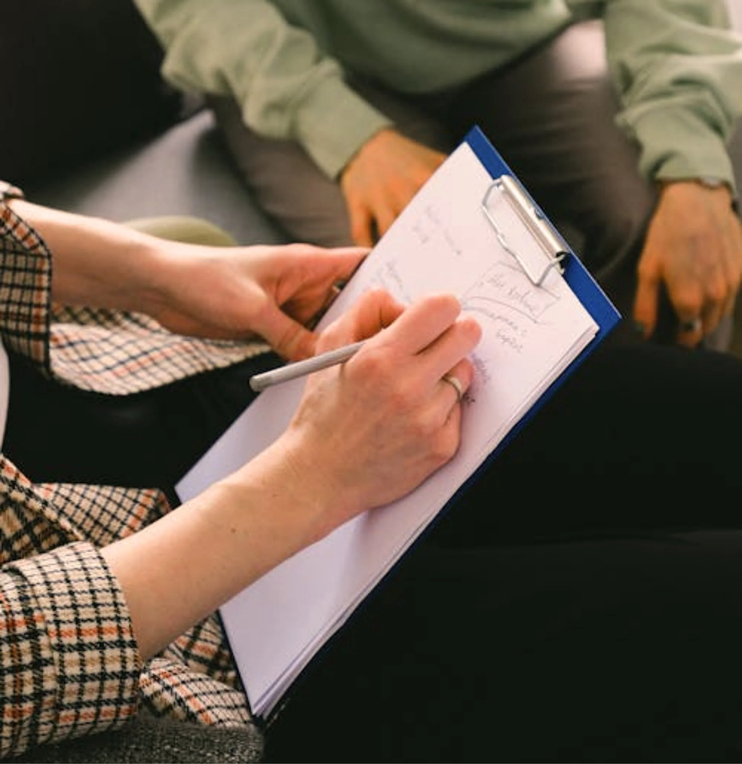 A person holding a clipboard and writing on a notepad, with another person sitting nearby.