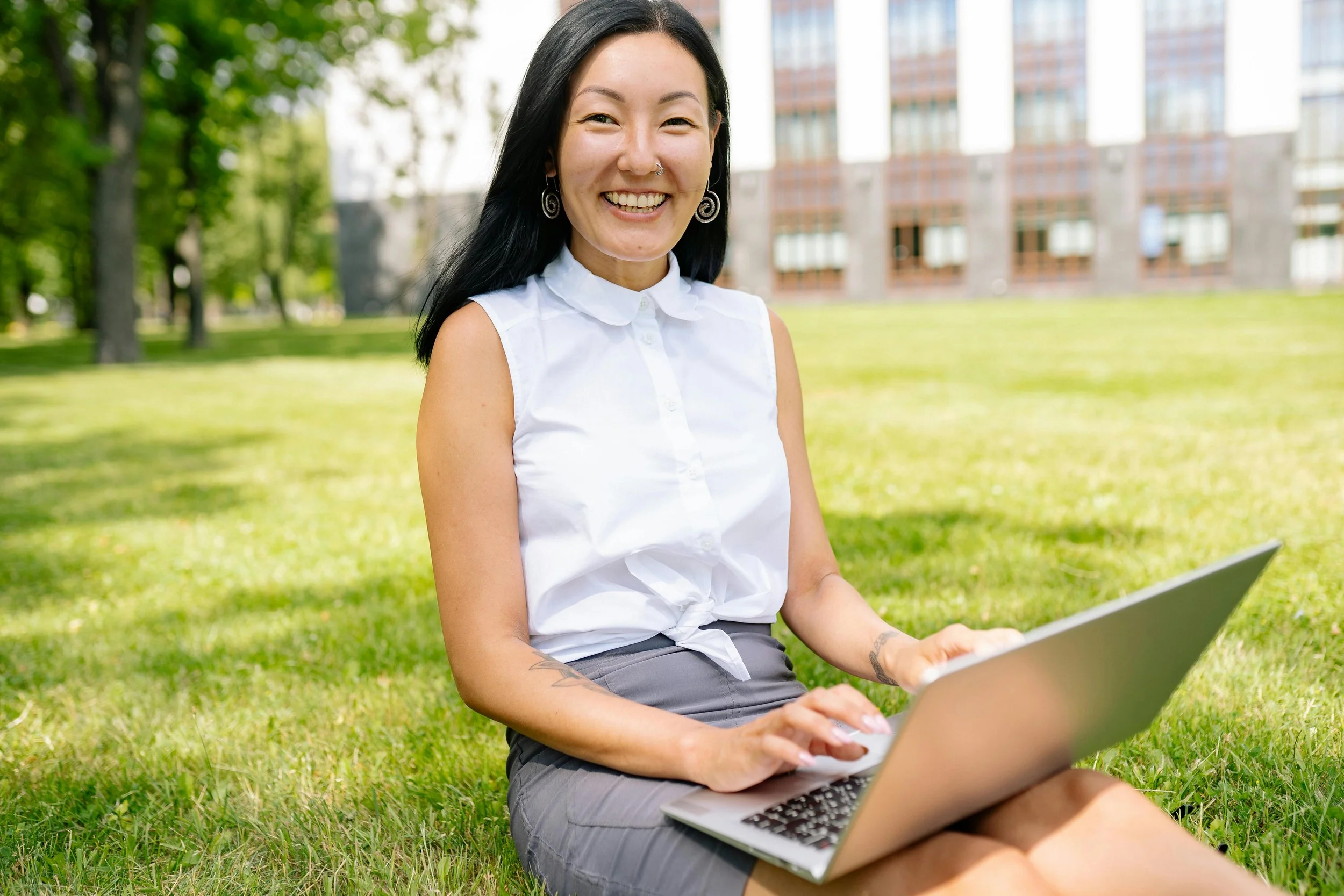 A young woman sitting on grass in a park, working on a laptop, smiling at the camera, with trees and a building in the background.