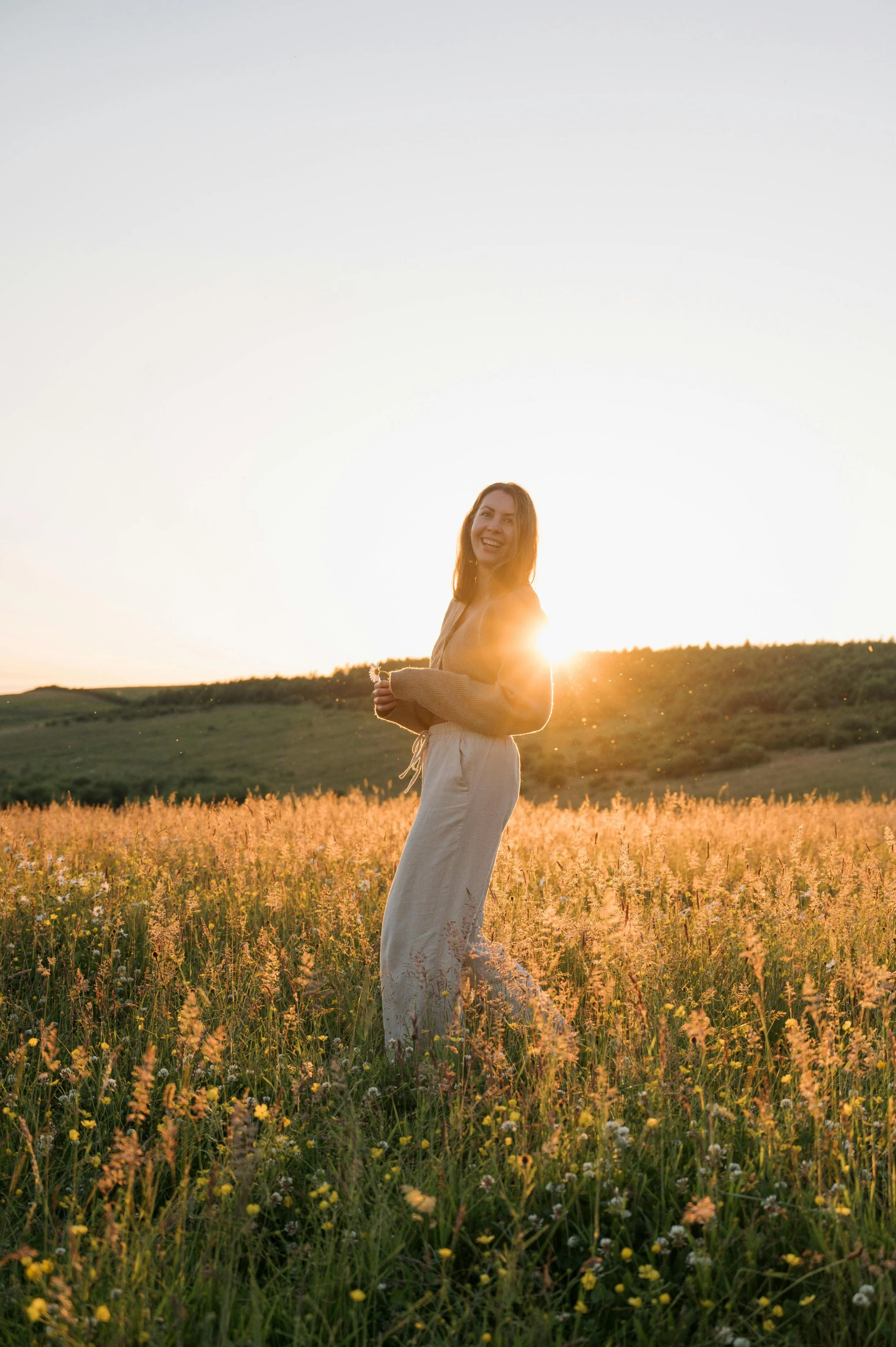 A woman standing in a field of tall grass and wildflowers during sunset, smiling and looking towards the camera with the sun behind her.