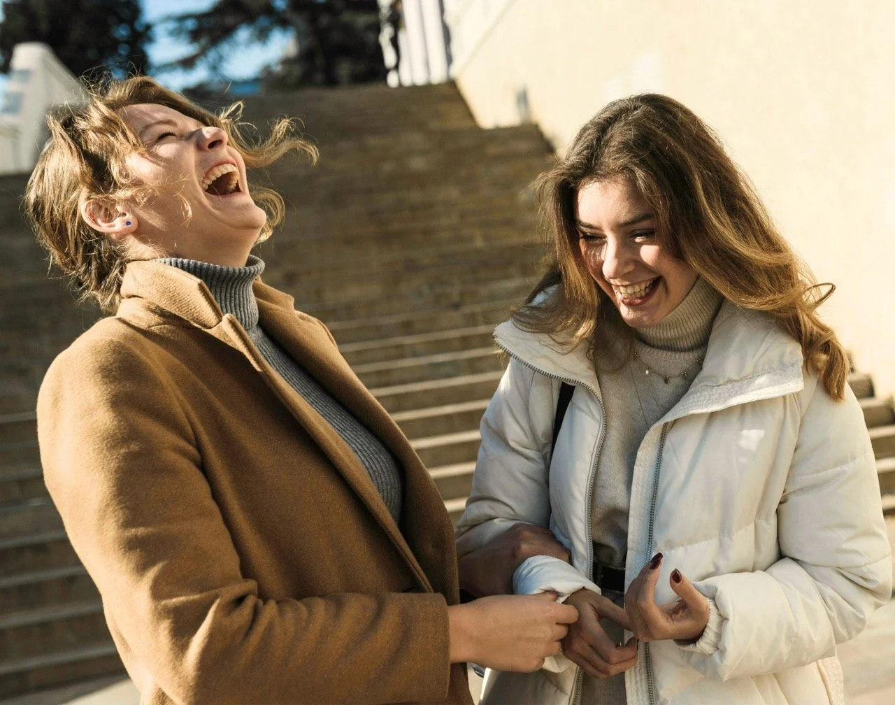 Two young women laughing and sharing a moment outdoors on steps, dressed in casual winter clothing.