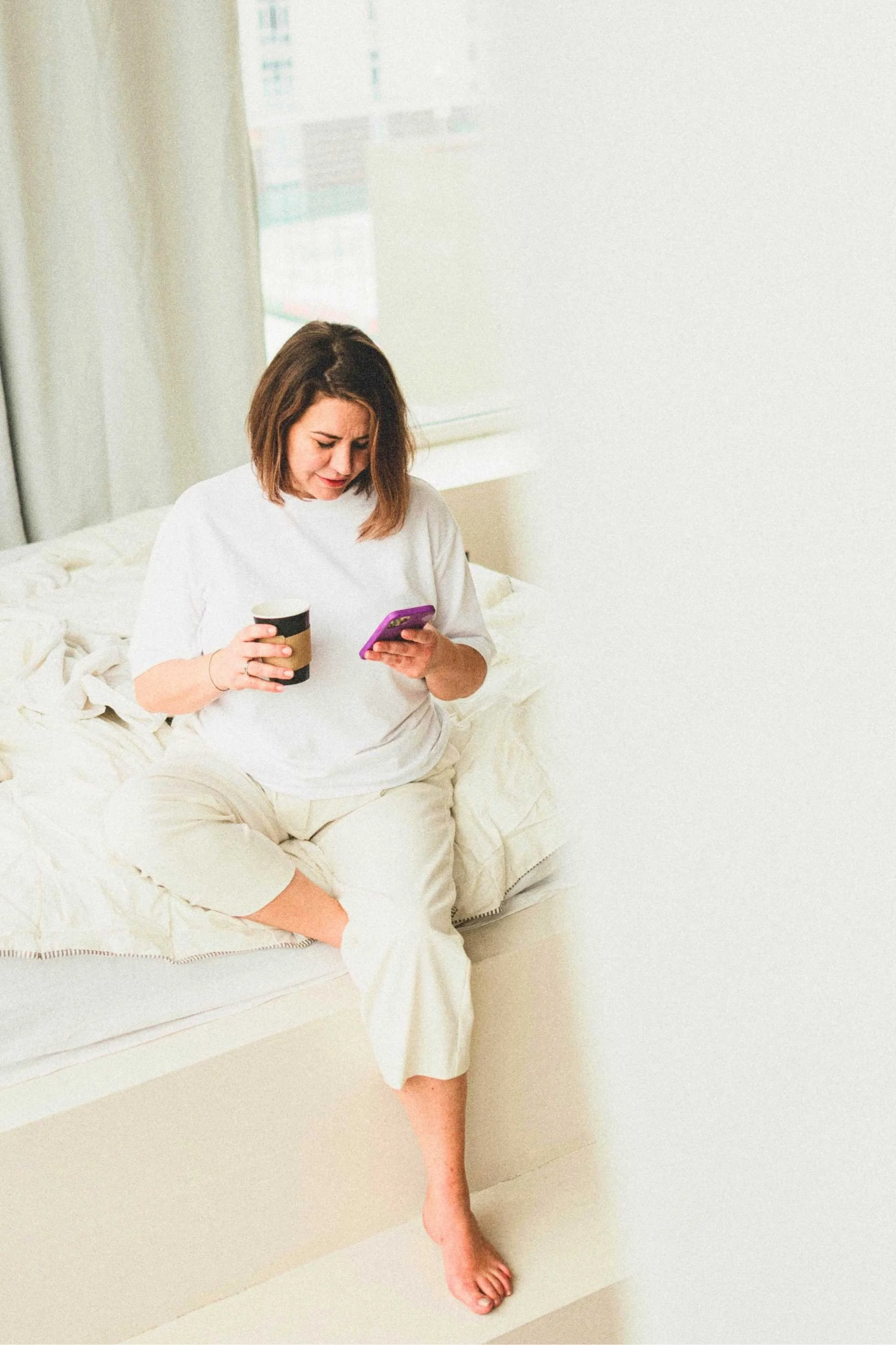 A woman sitting on a bed, looking at her phone, holding a coffee cup in her other hand, in a bright room with a large window.