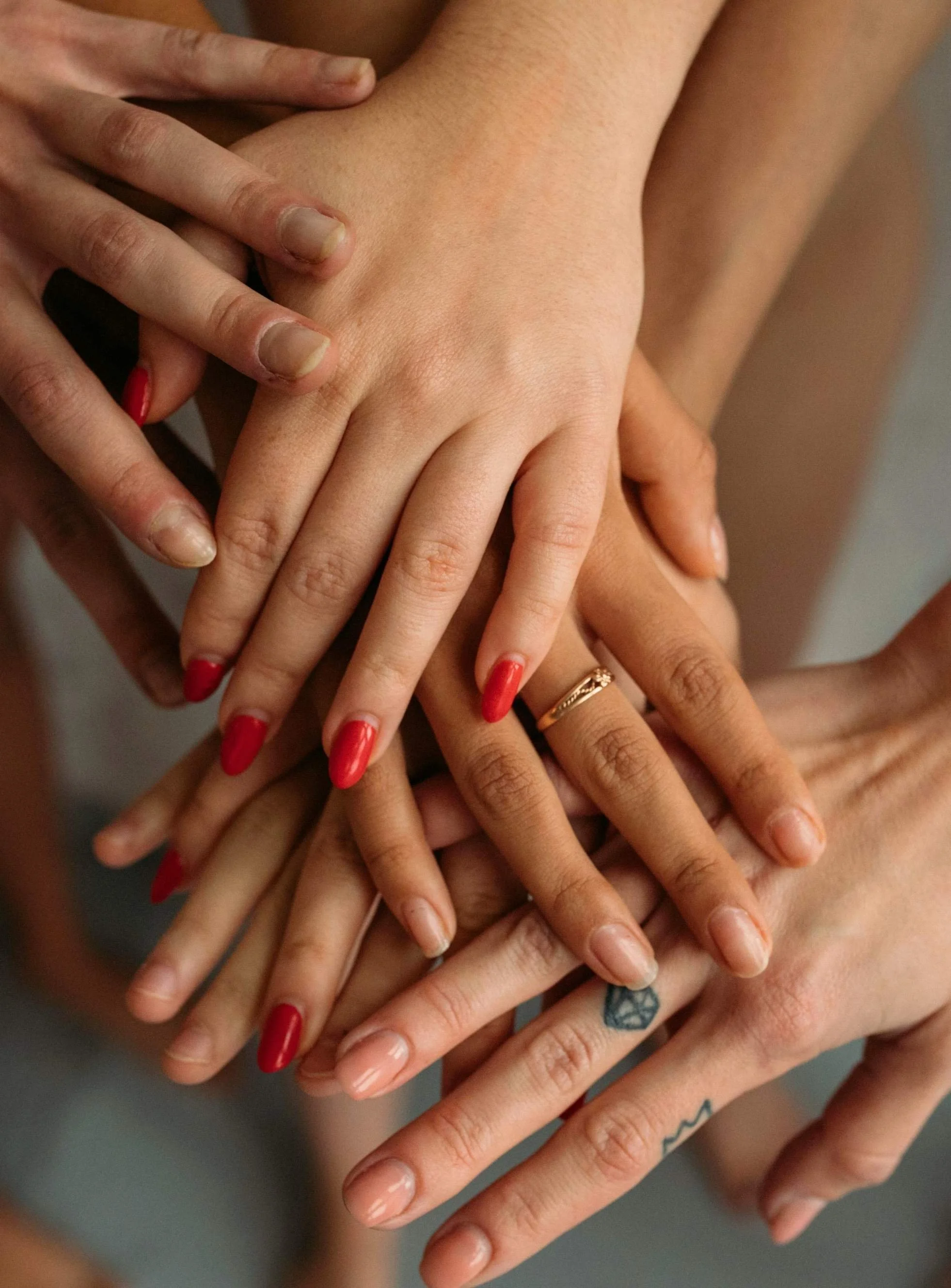 Multiple hands with diverse skin tones stacked together, some with tattoos and rings, and one with red nail polish, symbolizing unity and diversity.
