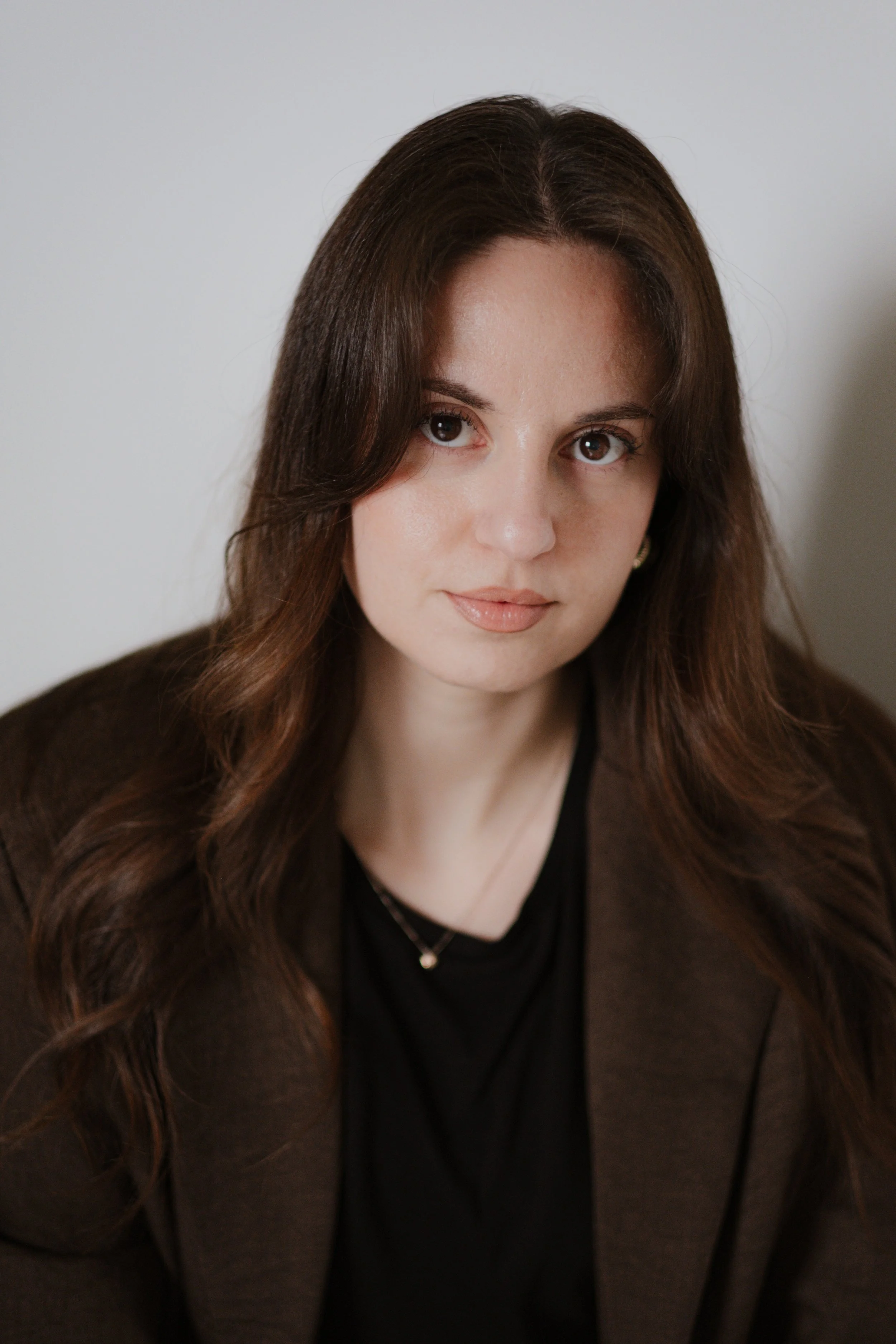 Close-up portrait of a young woman with long brown hair, wearing a black top and brown blazer, looking directly at the camera with a neutral expression.