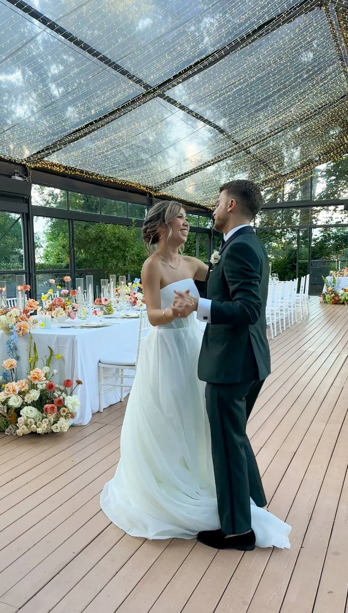 A bride and groom are dancing and smiling at their wedding reception, decorated with flowers and string lights.
