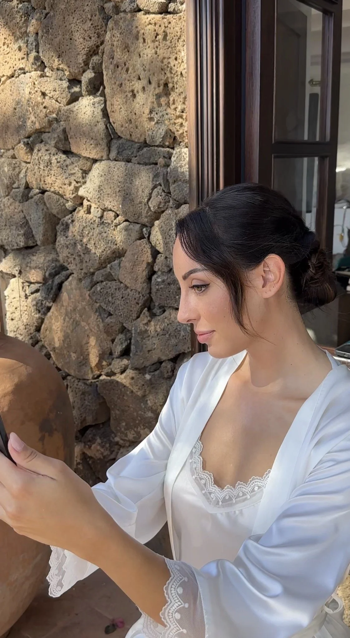 A woman with dark hair styled in an elegant updo, wearing a white satin robe with lace detail, looking at her phone while sitting outdoors next to a stone wall and a window.