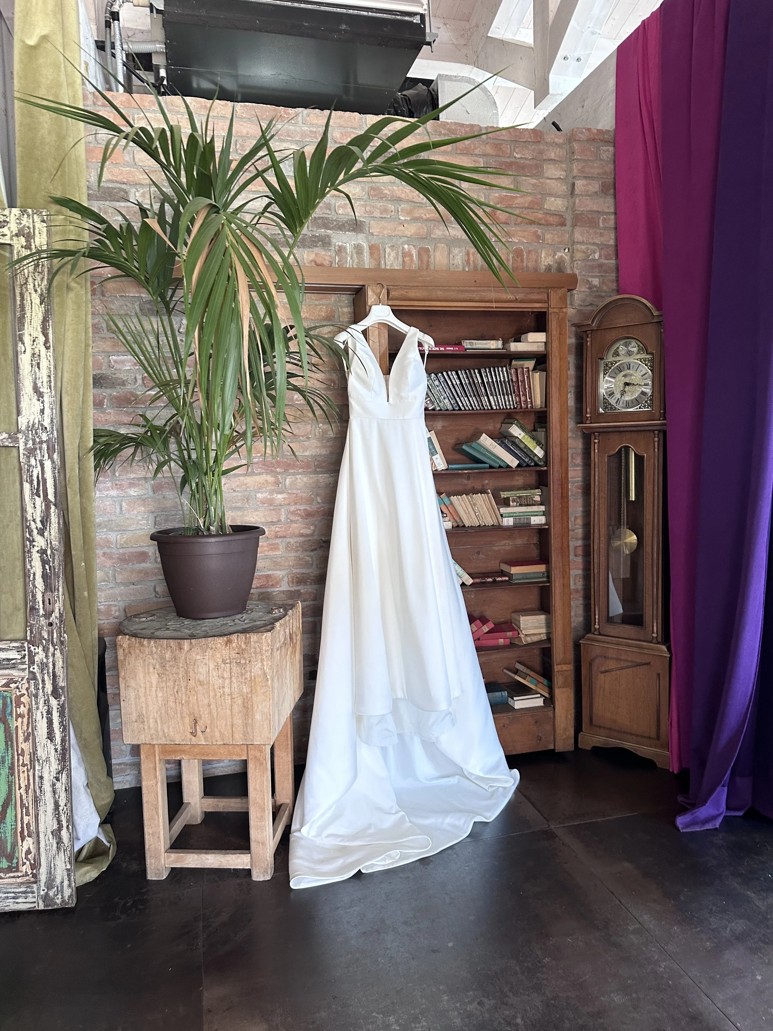 A white wedding dress hanging on a white hanger against a brick wall, with a potted plant, a wooden bookshelf filled with books, and an antique grandfather clock nearby.