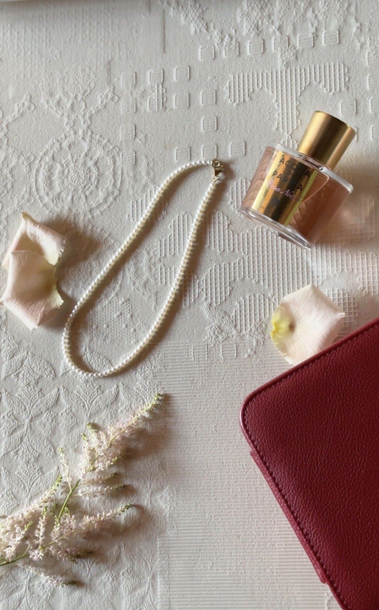 Flat lay of elegant accessories and personal items on a textured white surface, including a pearl necklace, pink rose petals and flower, a small bundle of dried flowers, a red leather wallet, a perfume bottle, and a pink-colored object.