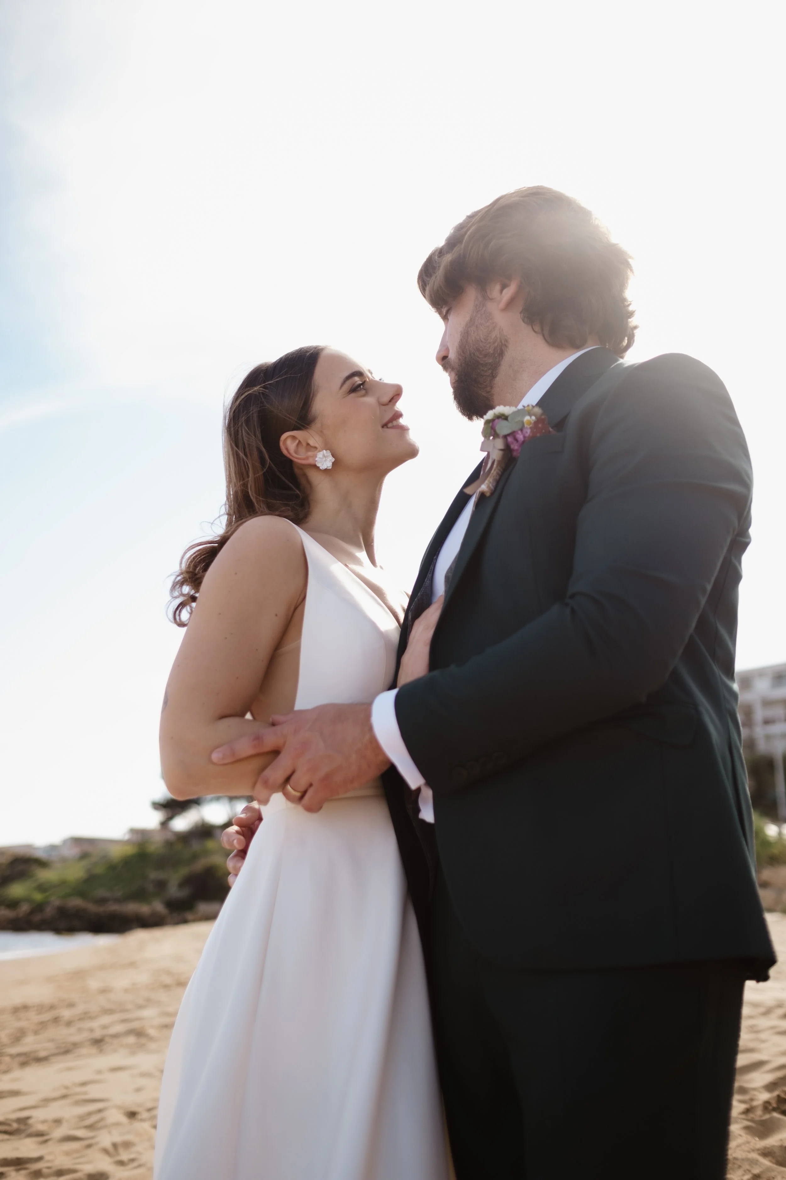 A bride and groom sharing a romantic moment on the beach, with the bride smiling at the groom who is looking at her, both dressed in wedding attire.
