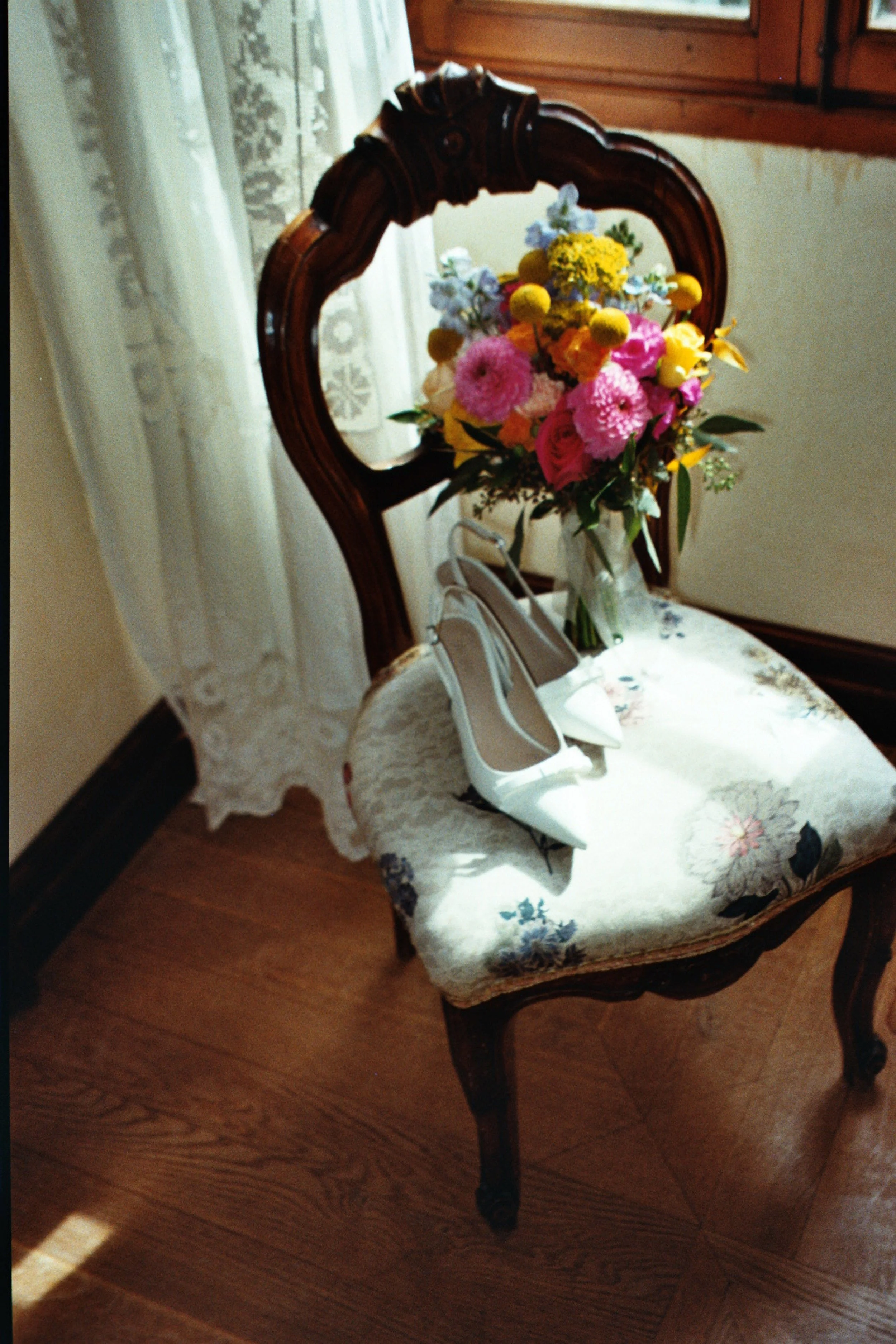 A vintage wooden chair with a floral patterned cushion, holding a pair of white high-heeled shoes, with a bouquet of colorful flowers on the backrest, placed on a wooden floor near a lace curtain and window.