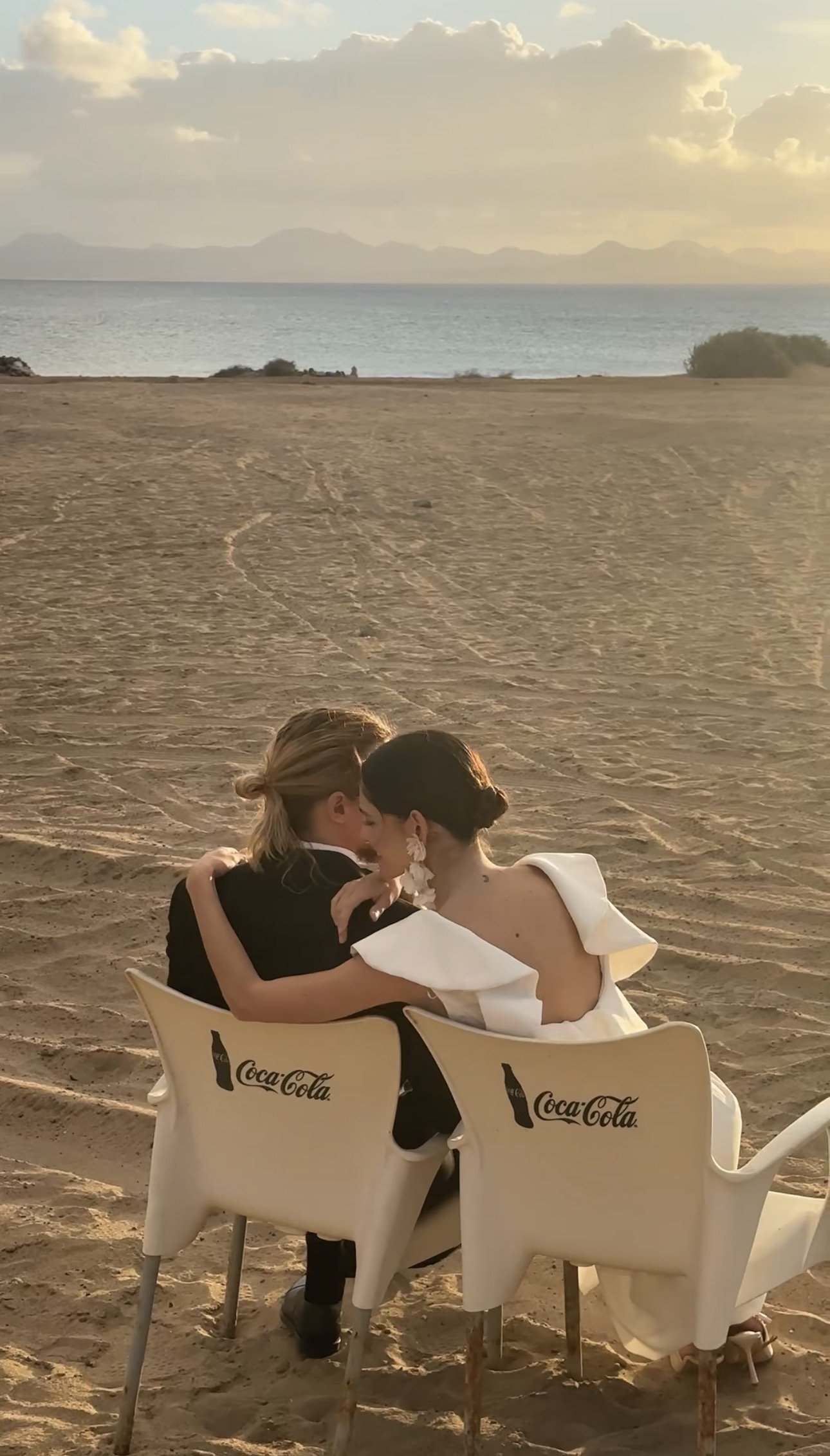 a me and a woman, dressed in wedding gowns, sitting closely together on beach chairs with Coca-Cola logos on the back, sharing an intimate moment at sunset overlooking the ocean.
