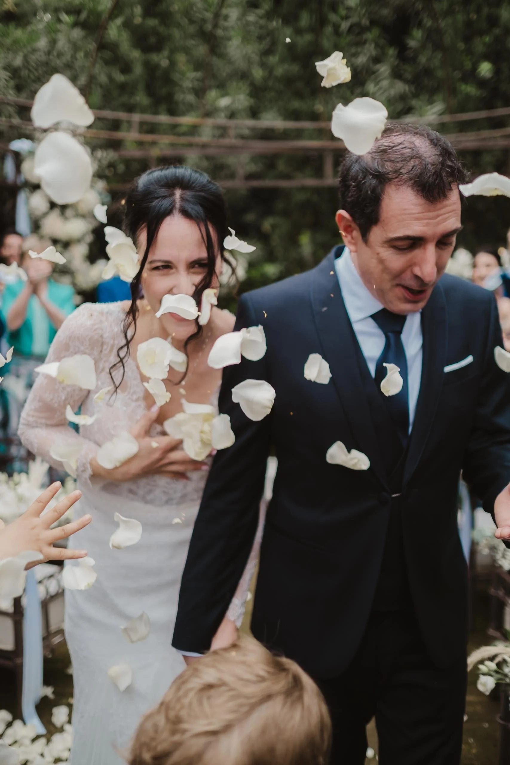 Wedding couple walking through shower of white flower petals, guests clapping in the background.