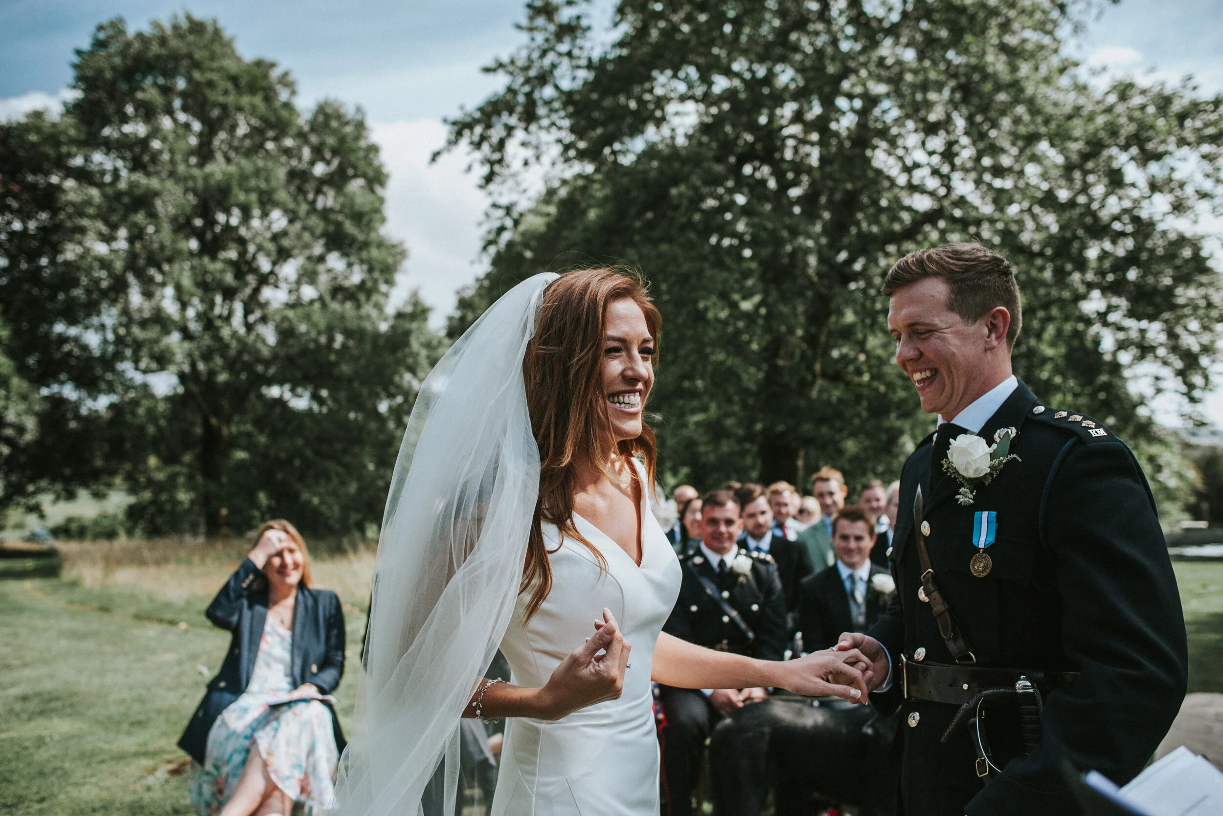 Bride and groom during wedding ceremony outdoors, holding hands, smiling, with guests seated behind them on a grassy area with trees.