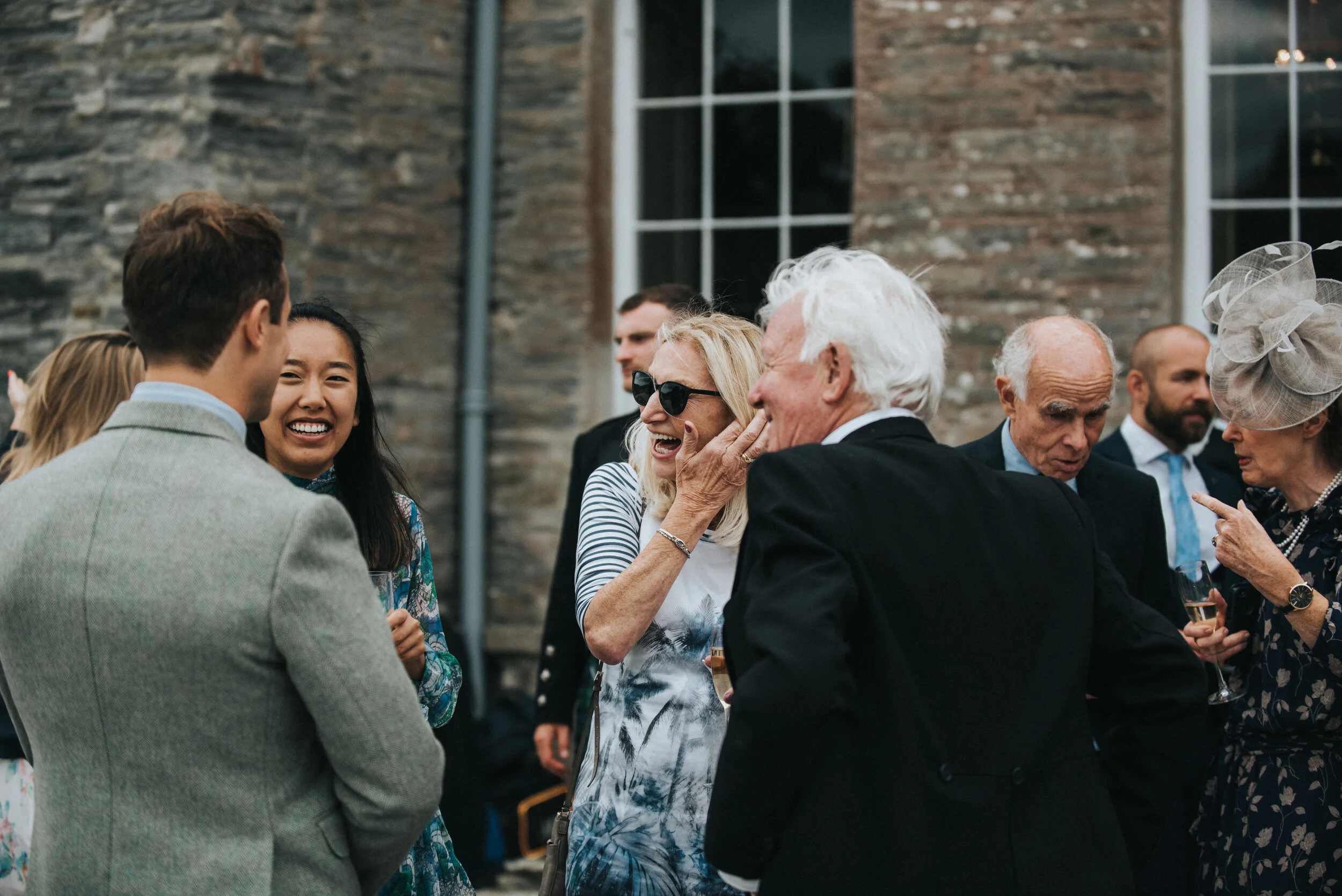 Group of people at an outdoor social event, talking and laughing, with a brick building in the background.
