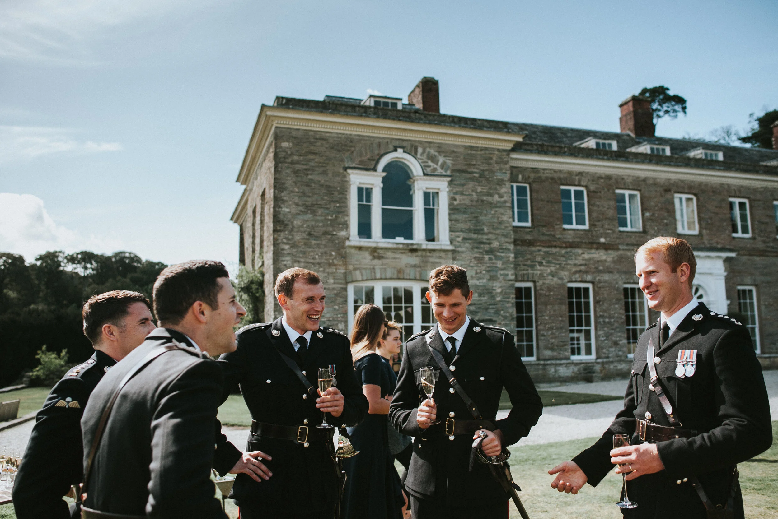 Group of men in formal military dress uniform holding champagne glasses and chatting outside a large stone mansion.