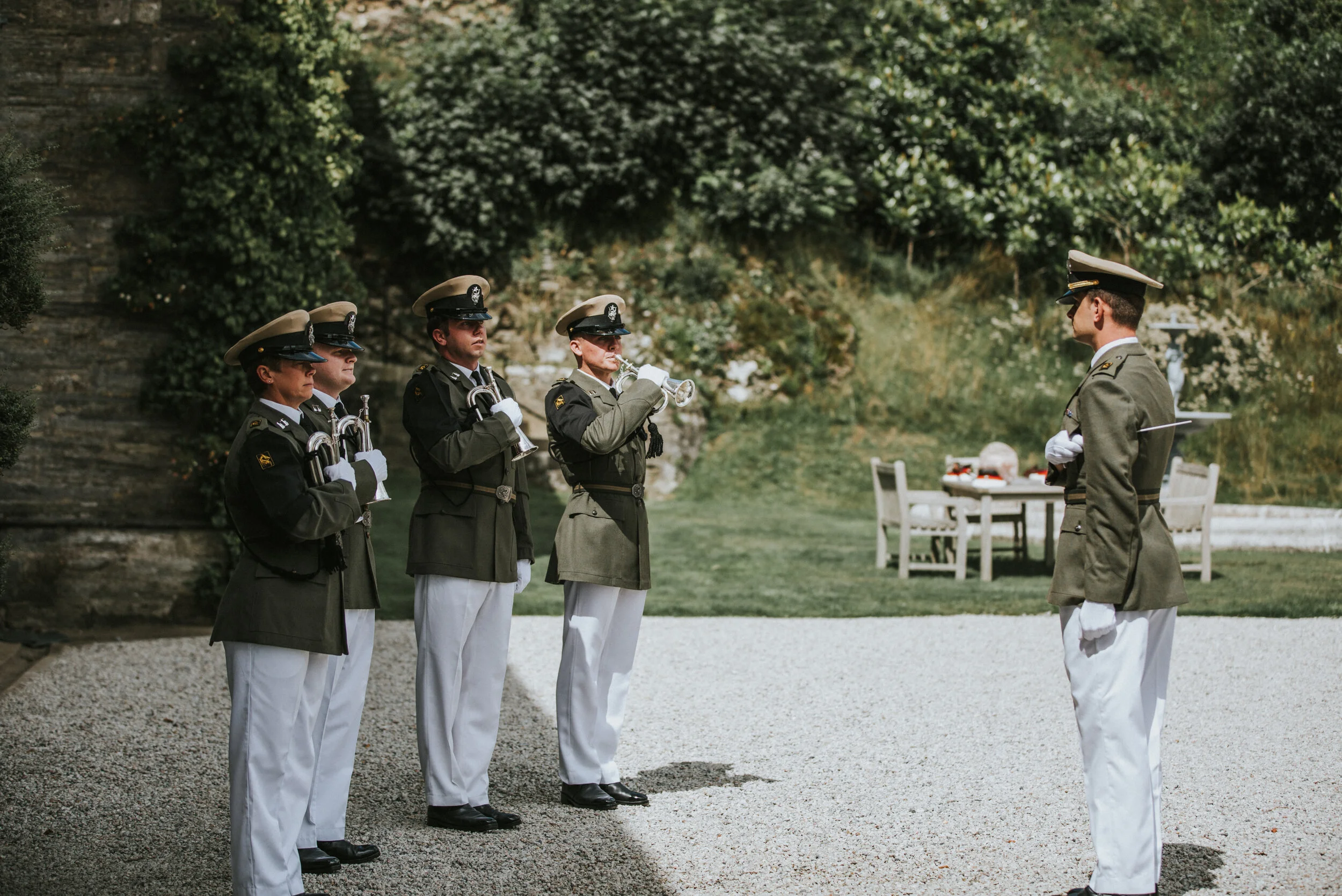 Military ceremony with four soldiers standing in a line, playing trumpets, facing a commanding officer in an outdoor setting with greenery and a table in the background.