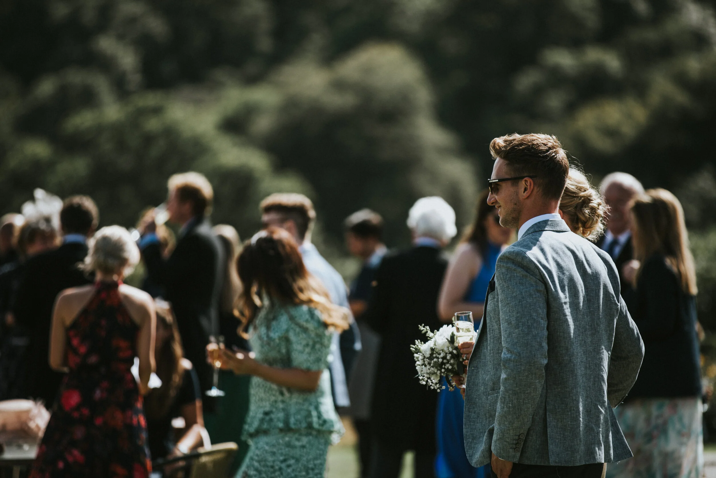 A man in a gray suit with sunglasses holding a glass of champagne and a bouquet, at an outdoor wedding reception with guests in formal attire in the background.