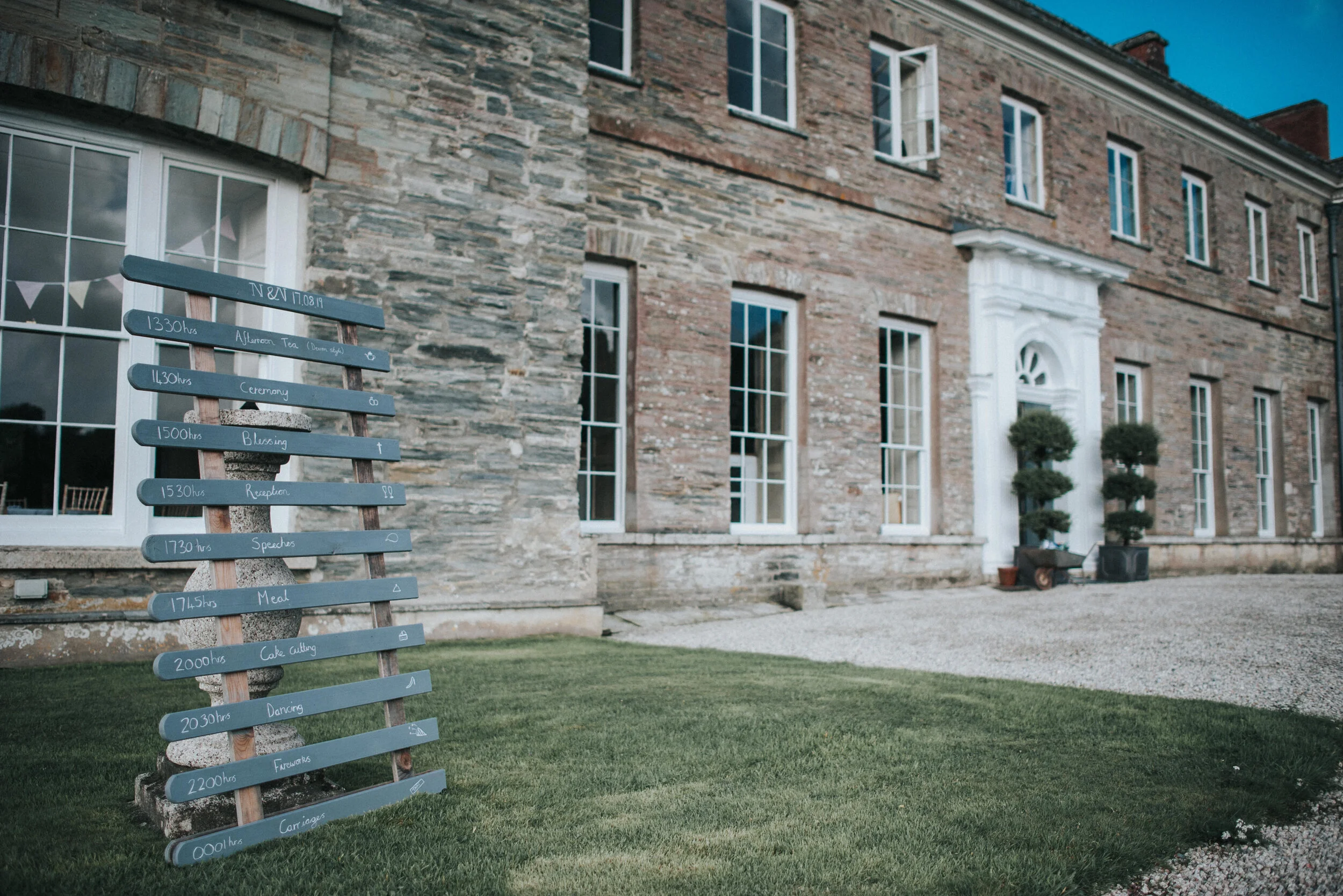 A wooden schedule board with handwritten event times and titles, positioned on a lawn outside a brick mansion with tall windows and two potted topiary trees.