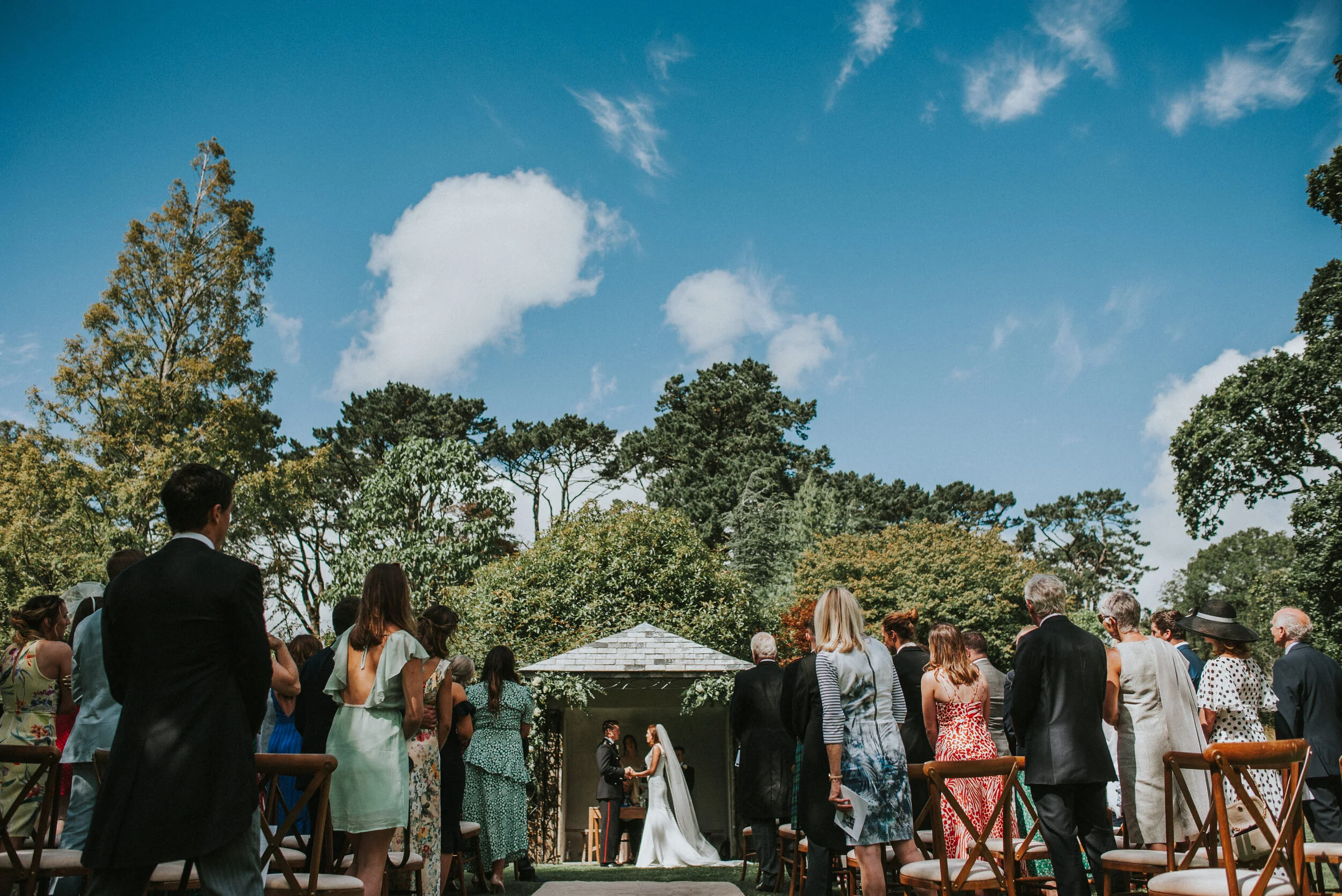 Outdoor wedding ceremony with guests standing and facing an officiant and bride and groom under a pavilion, surrounded by trees and a blue sky with clouds.