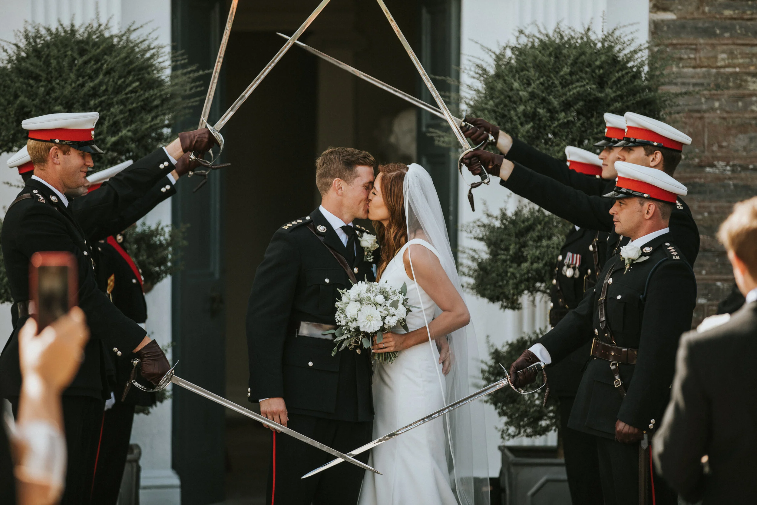 A wedding couple, in military dress, sharing a kiss under a sword arch made by uniformed men in dress uniforms with red and white hats.