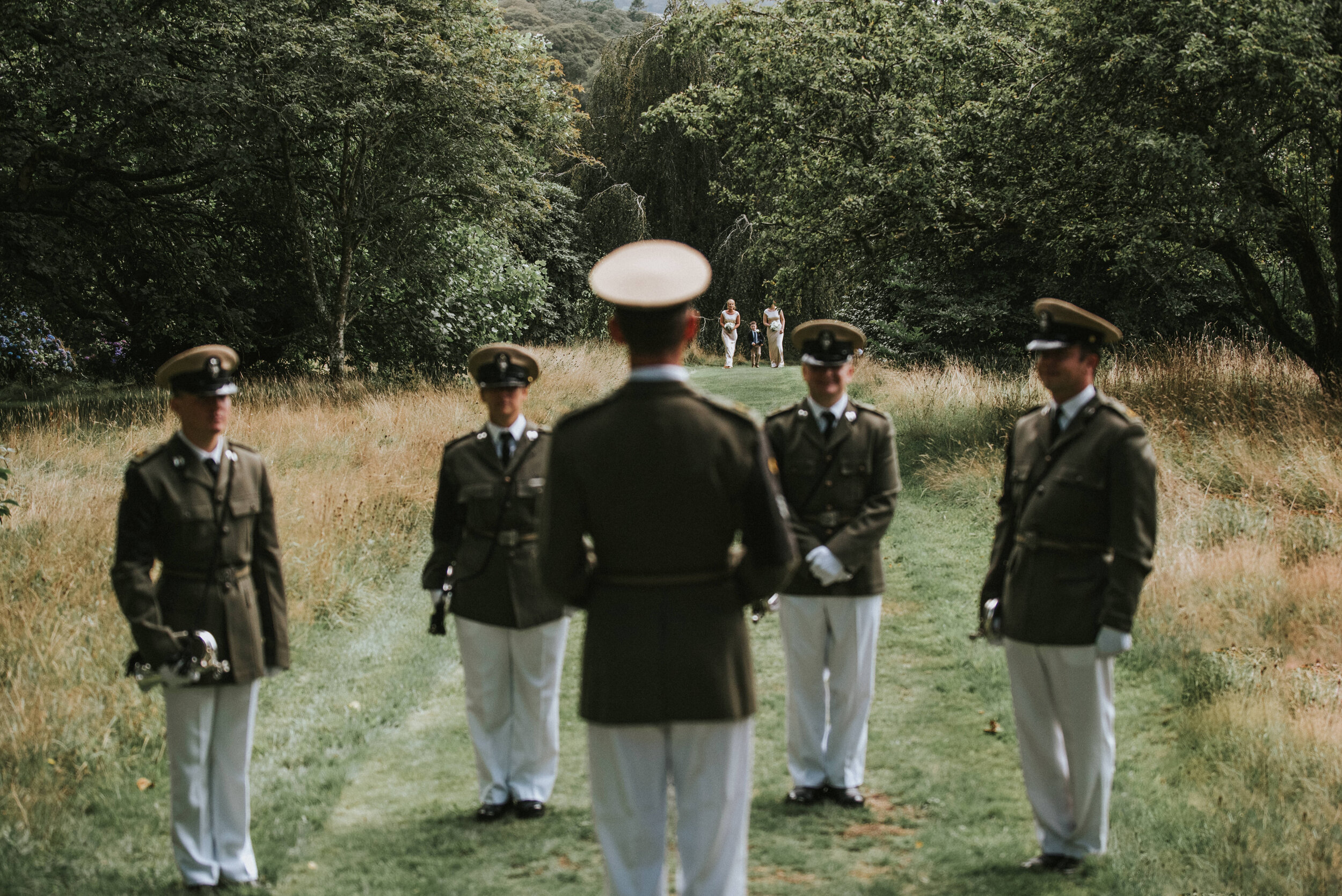 Military officers in uniform standing in a row on a grassy path in a park, with a group of women standing in the background at the end of the path. The scene is outdoors with trees surrounding the area.