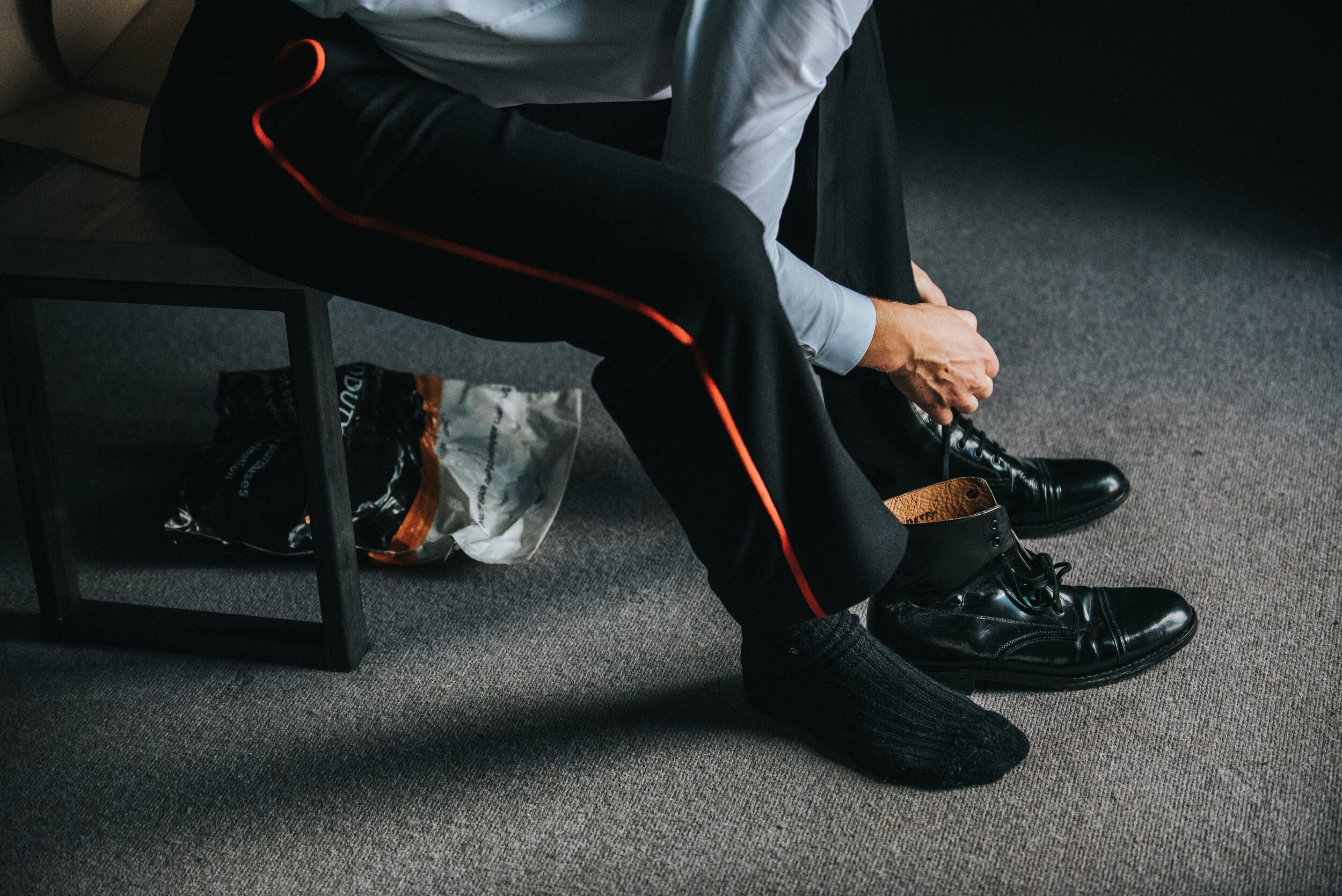 A man in business attire is sitting on a chair, tying his shoe. He is wearing black dress shoes, black pants with a red stripe, and a light-colored shirt. There is a plastic bag with snacks or food underneath the chair.