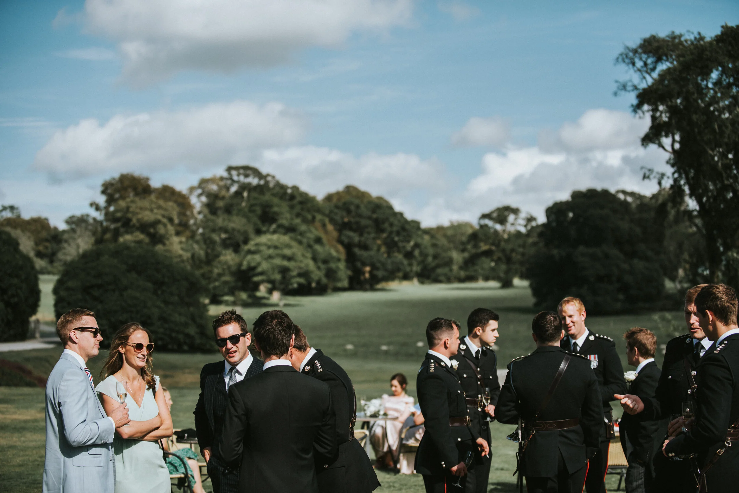People in formal attire, including military uniforms, socializing outdoors on a grassy field under a partly cloudy sky.