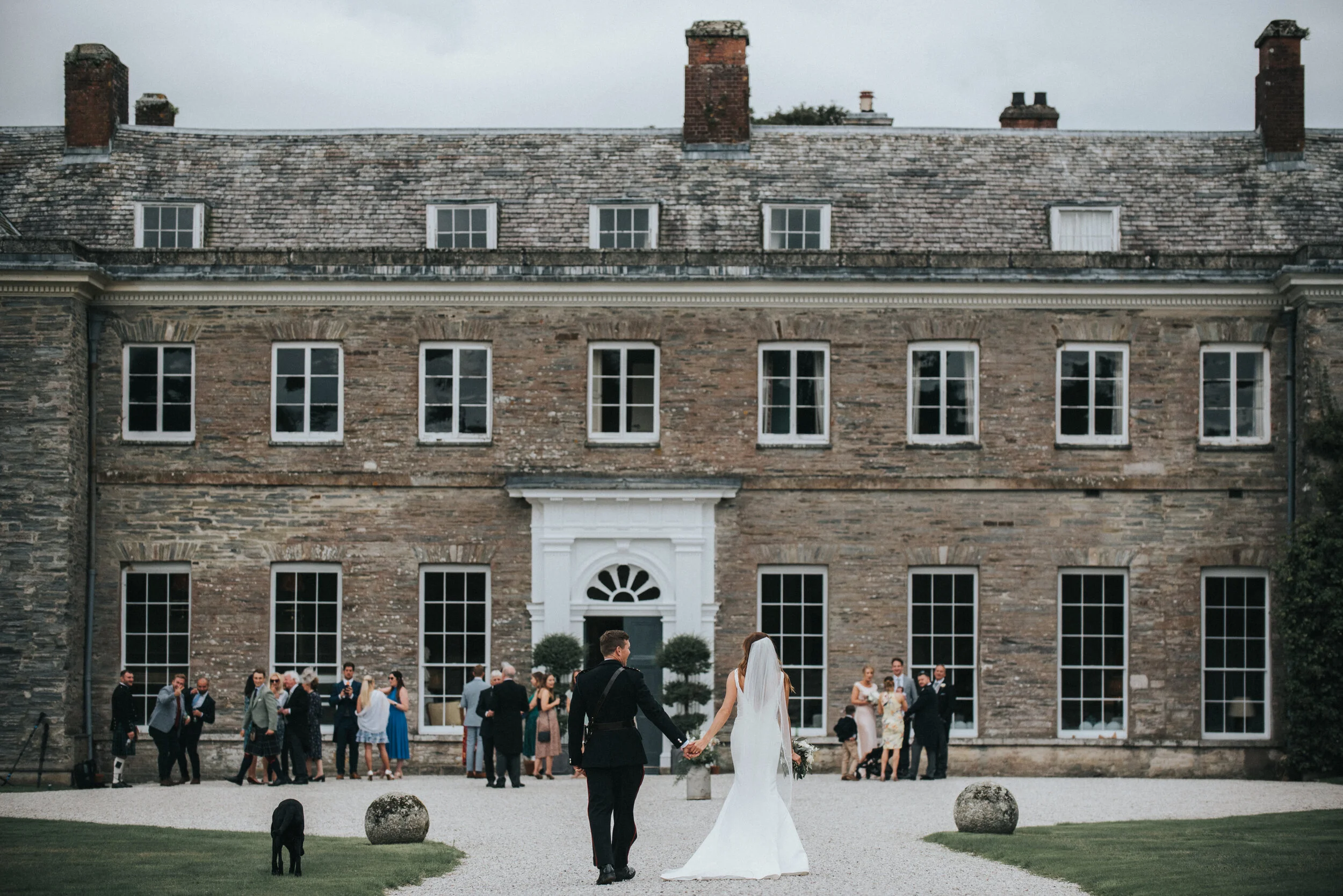 A newlywed couple holding hands walks away from a large historic brick building with many windows, while wedding guests gather in the background.