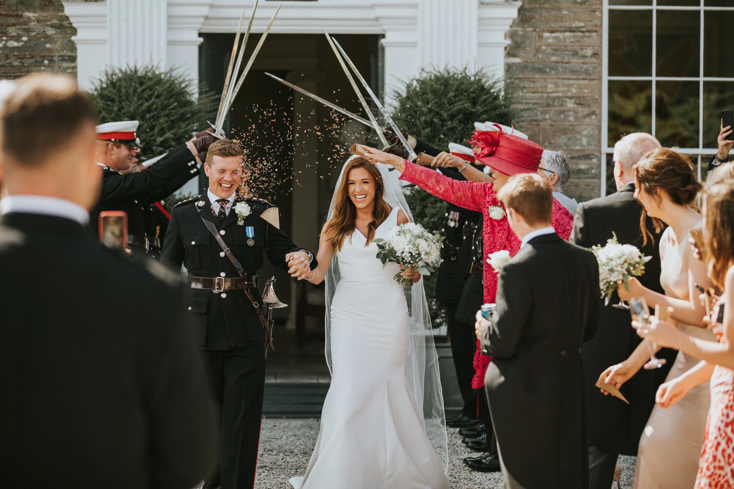 Bride and groom smiling as they walk through a crowd of wedding guests who are celebrating with confetti and sword arch entrance outside a building.