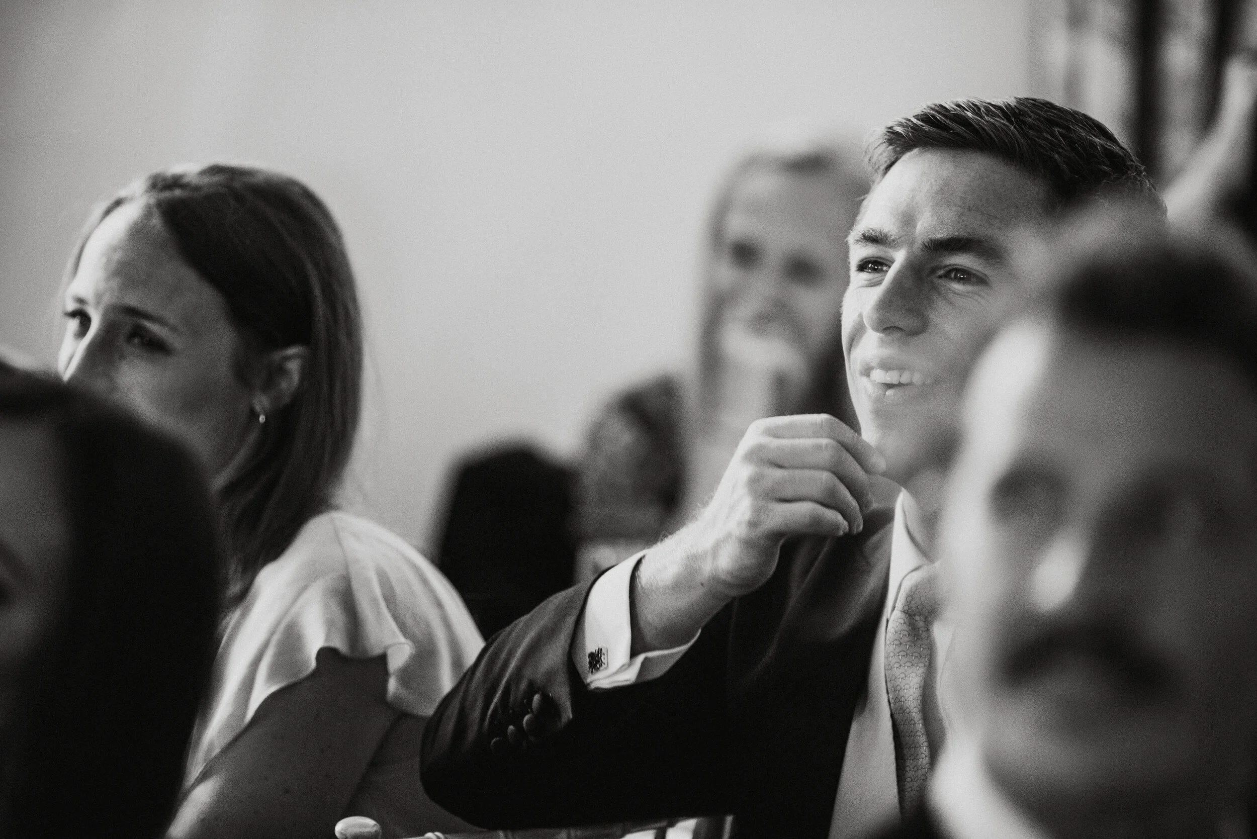 Black and white photo of a group of people attending an event, with a young man in a suit smiling and resting his chin on his hand in focus.