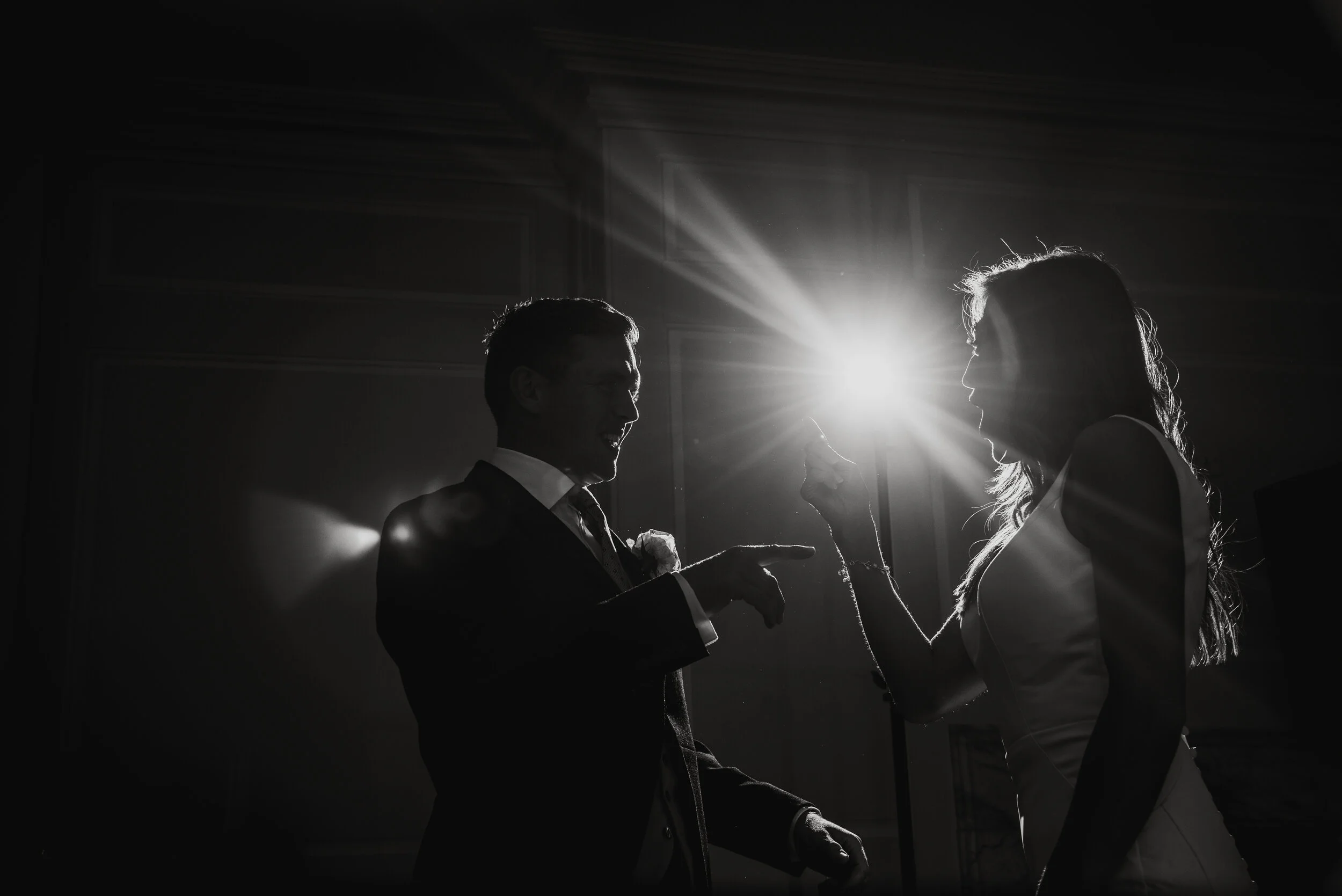 Silhouetted man and woman dancing and laughing at a wedding reception, backlit with bright light creating a dramatic scene.