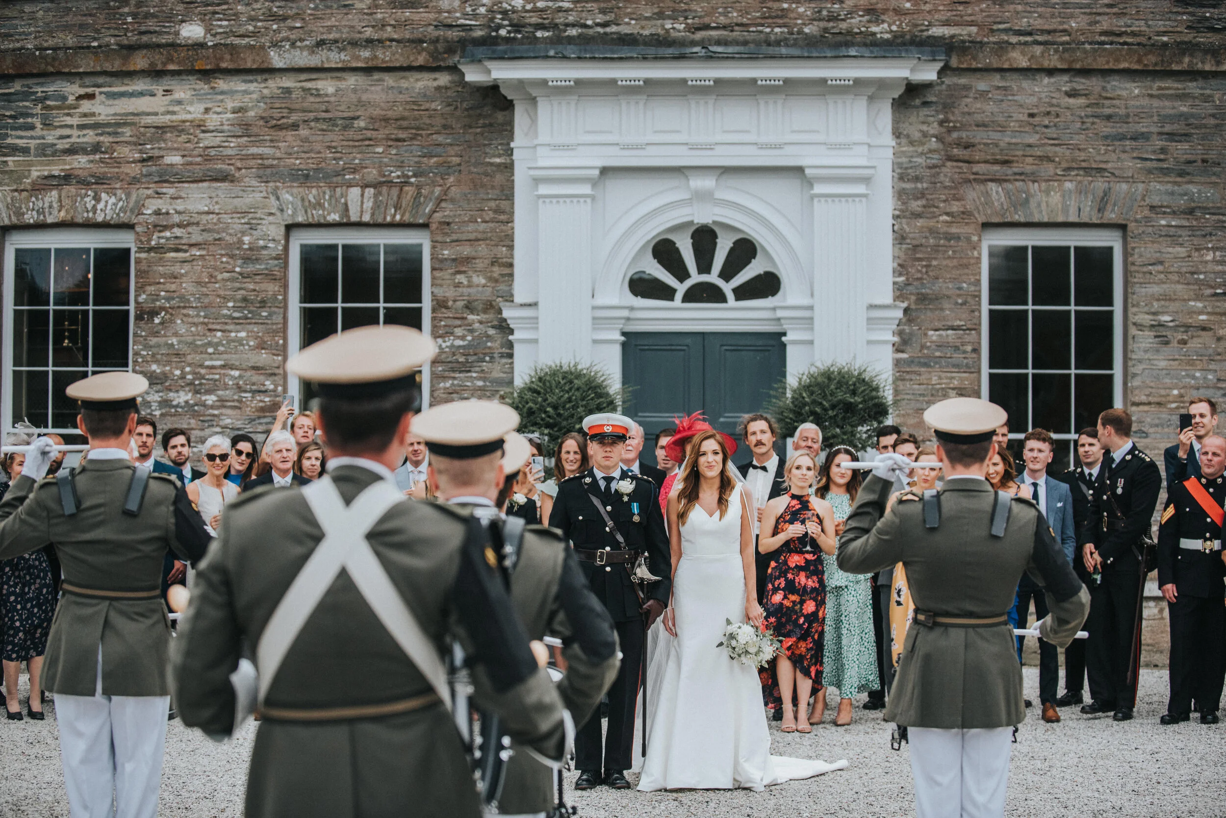 A wedding ceremony with military personnel honor guard in front of a brick building with a white stone entryway, crowds of guests watching.