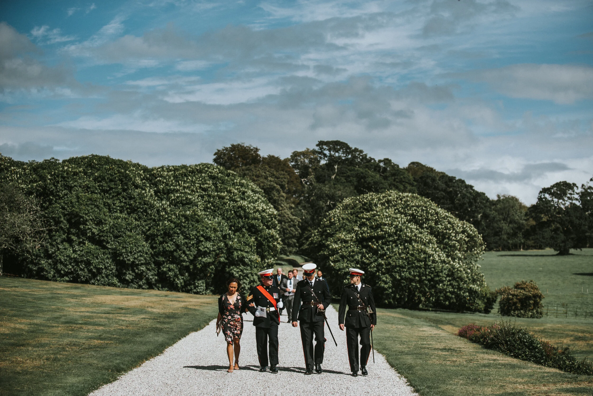 Group of military personnel walking on a gravel path with a woman in a floral dress in a park-like setting with lush green trees and cloudy sky.