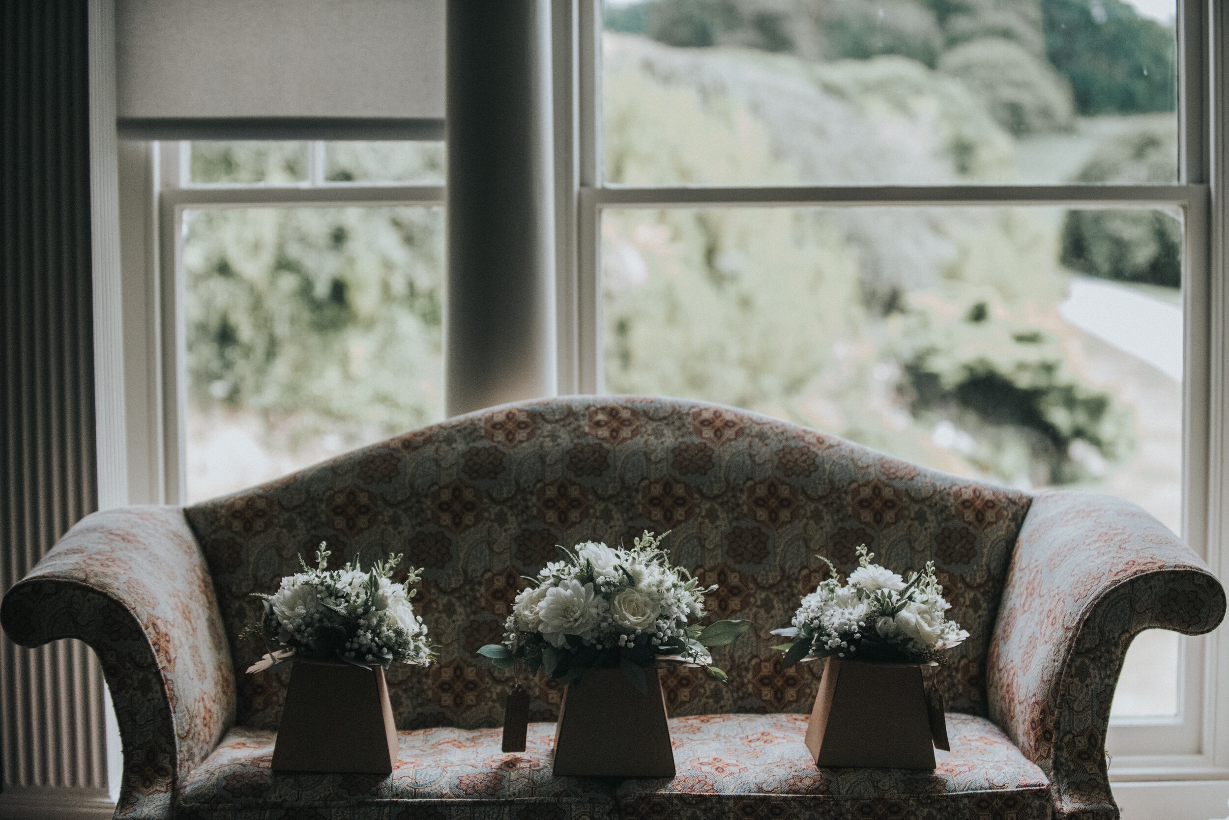 A vintage upholstered sofa with floral fabric, placed in front of a large window, holding three arrangements of white flowers in vases.