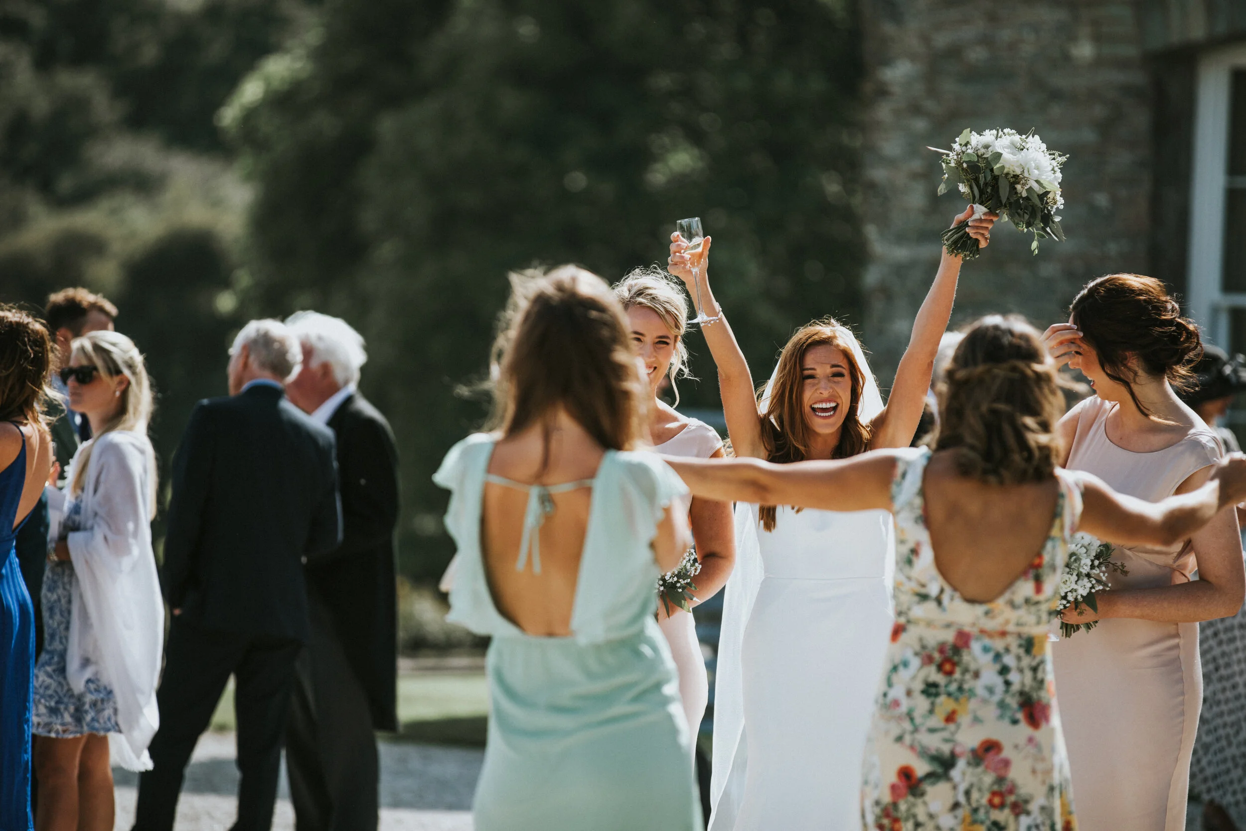 Group of women celebrating outdoors at a wedding, with one woman holding a bouquet of flowers and another raising a glass.