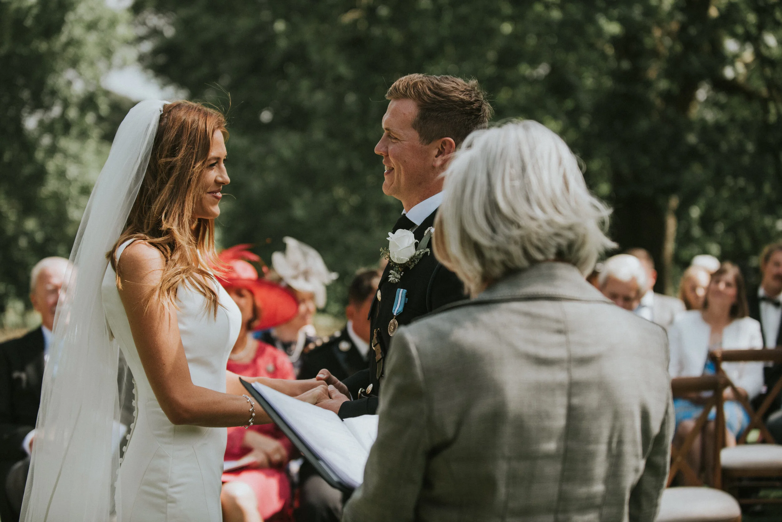 Bride and groom exchanging vows during an outdoor wedding ceremony, with guests seated in the background.