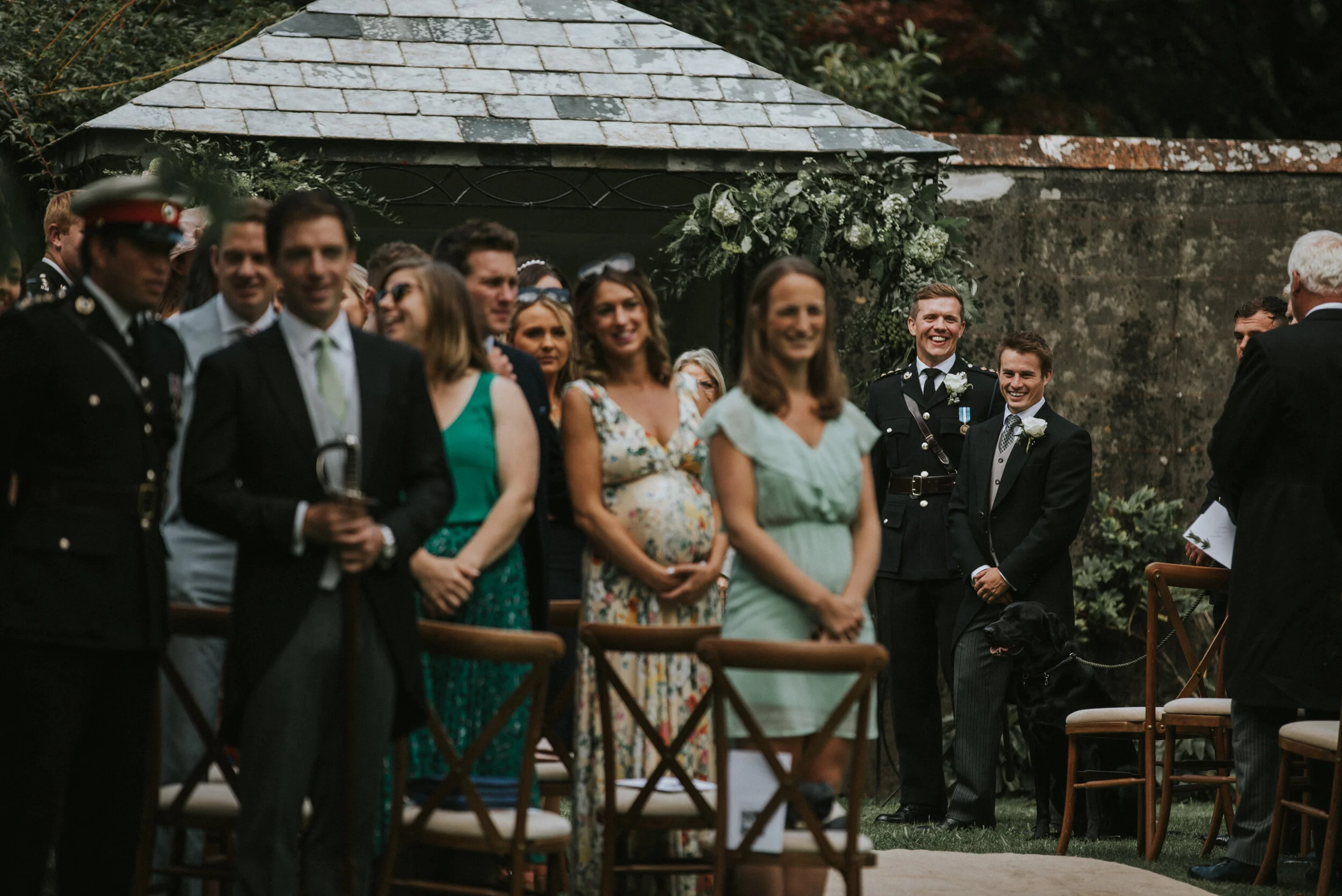 People gathered outdoors at a wedding ceremony, some are smiling and looking towards the officiant, with chairs and greenery around.