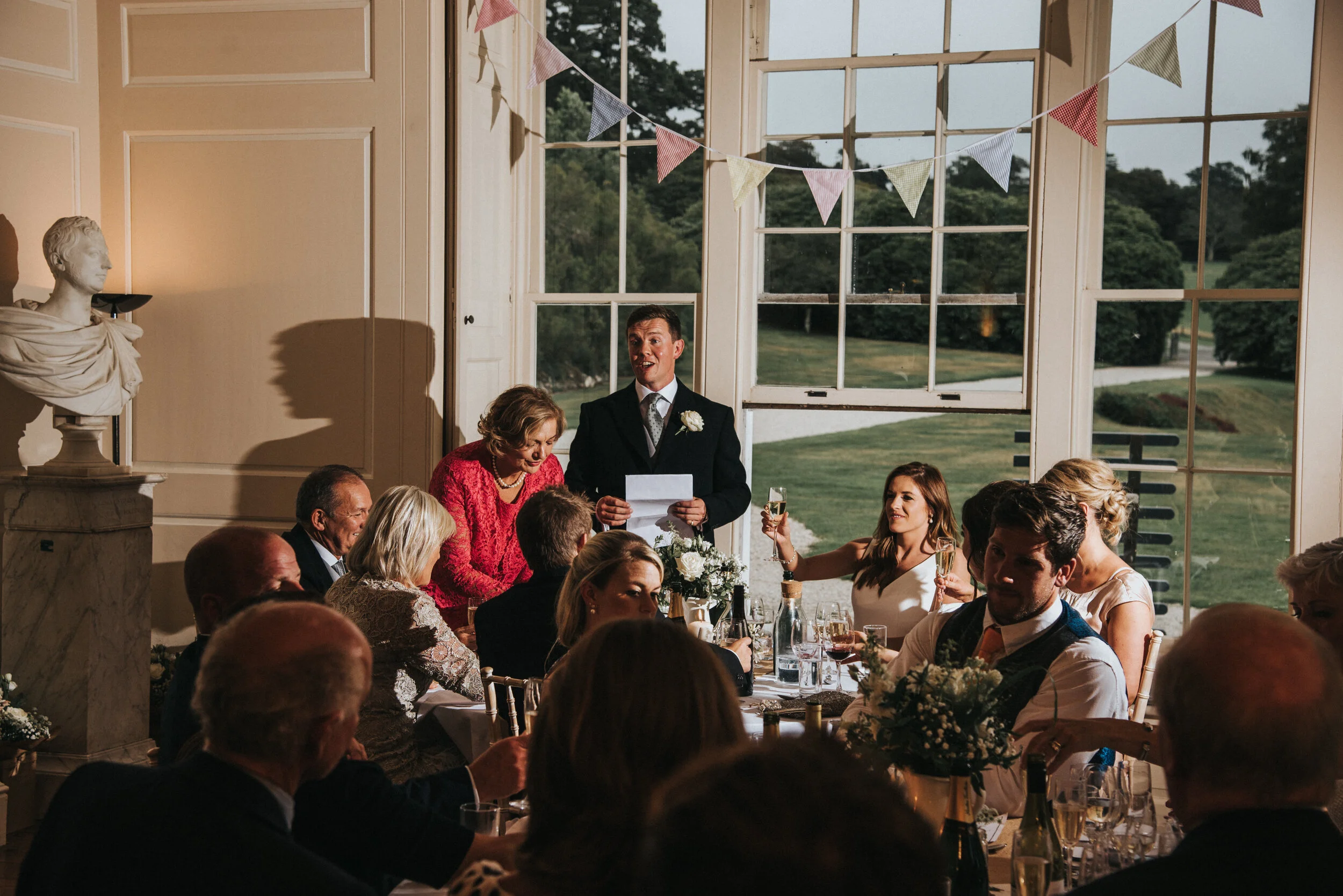 At a wedding reception, a man in a suit is giving a speech while holding a piece of paper. Guests are seated at a decorated table, raising glasses in toast. Large windows reveal a green outdoor landscape, and festive bunting is hung across the room.