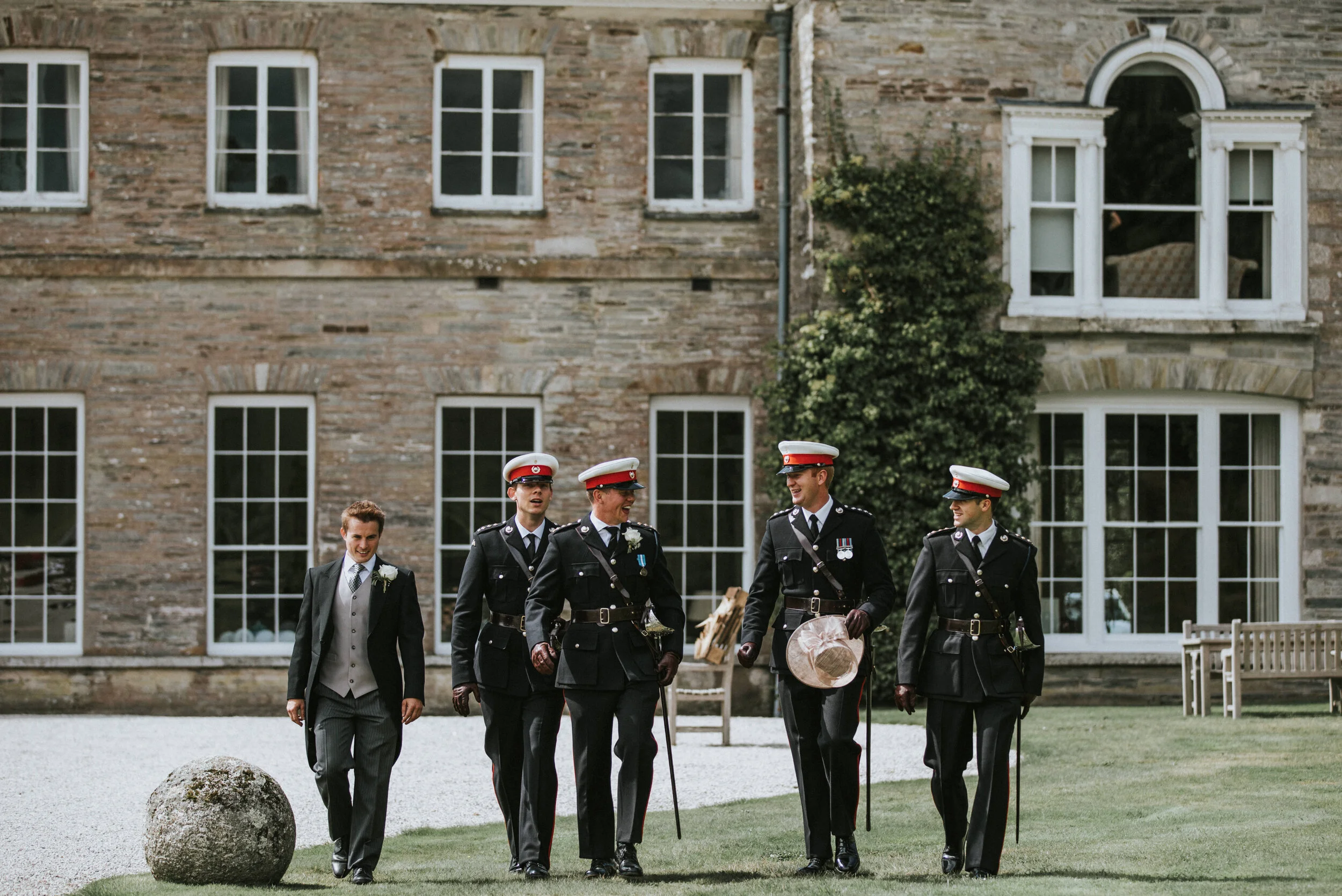 Five men, including a groom and four officers in uniform, walking together outdoors on a green lawn with a stone building in the background.
