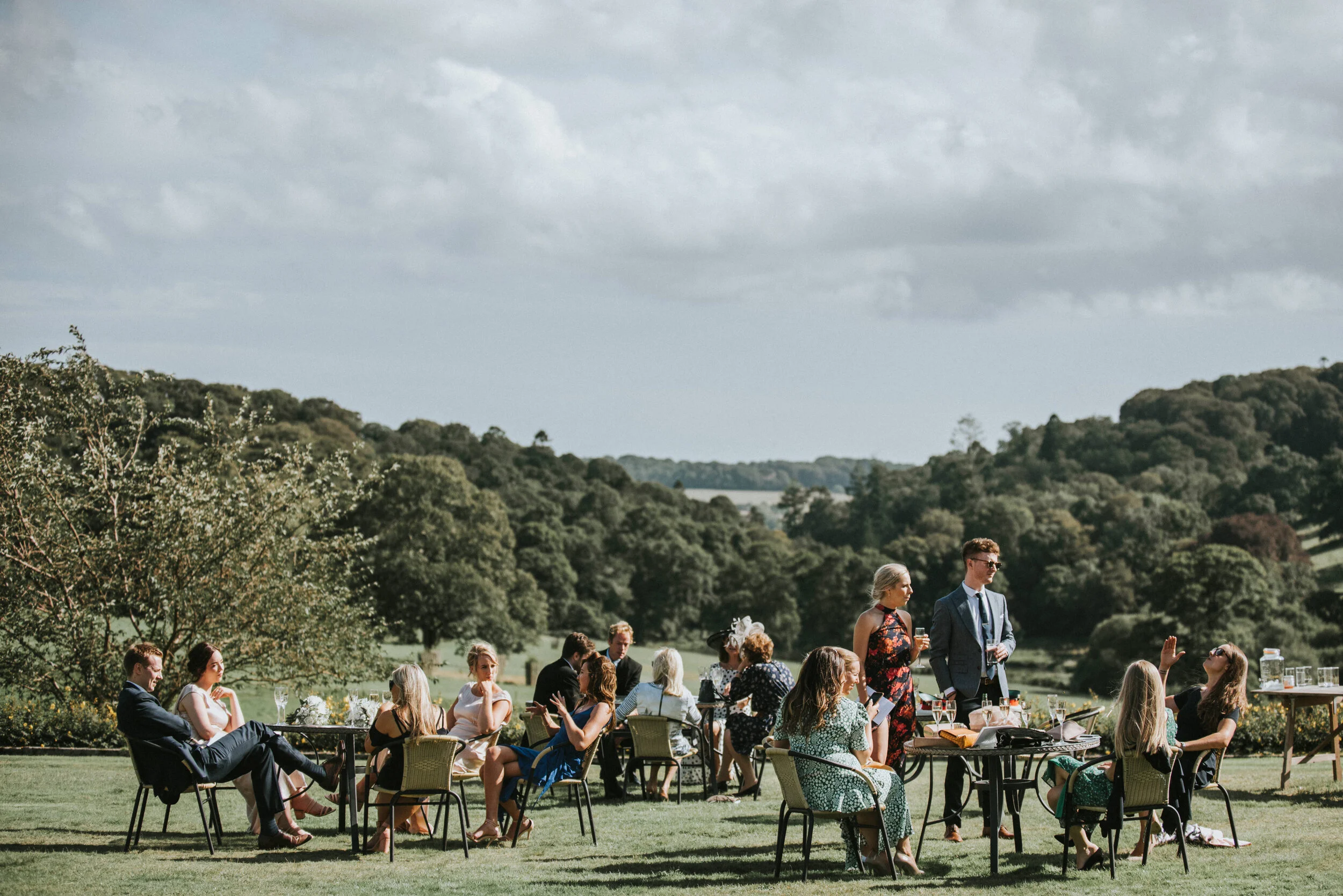 People gathered outdoors on a grassy area with trees and hills in the background, sitting at tables and standing, socializing at a daytime event.