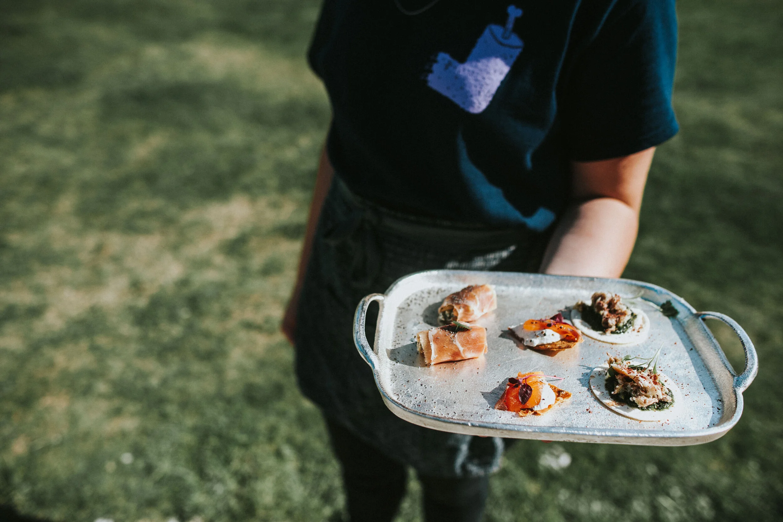 A person holding a silver tray with six small appetizers, including crostinis, on a grassy outdoor setting.