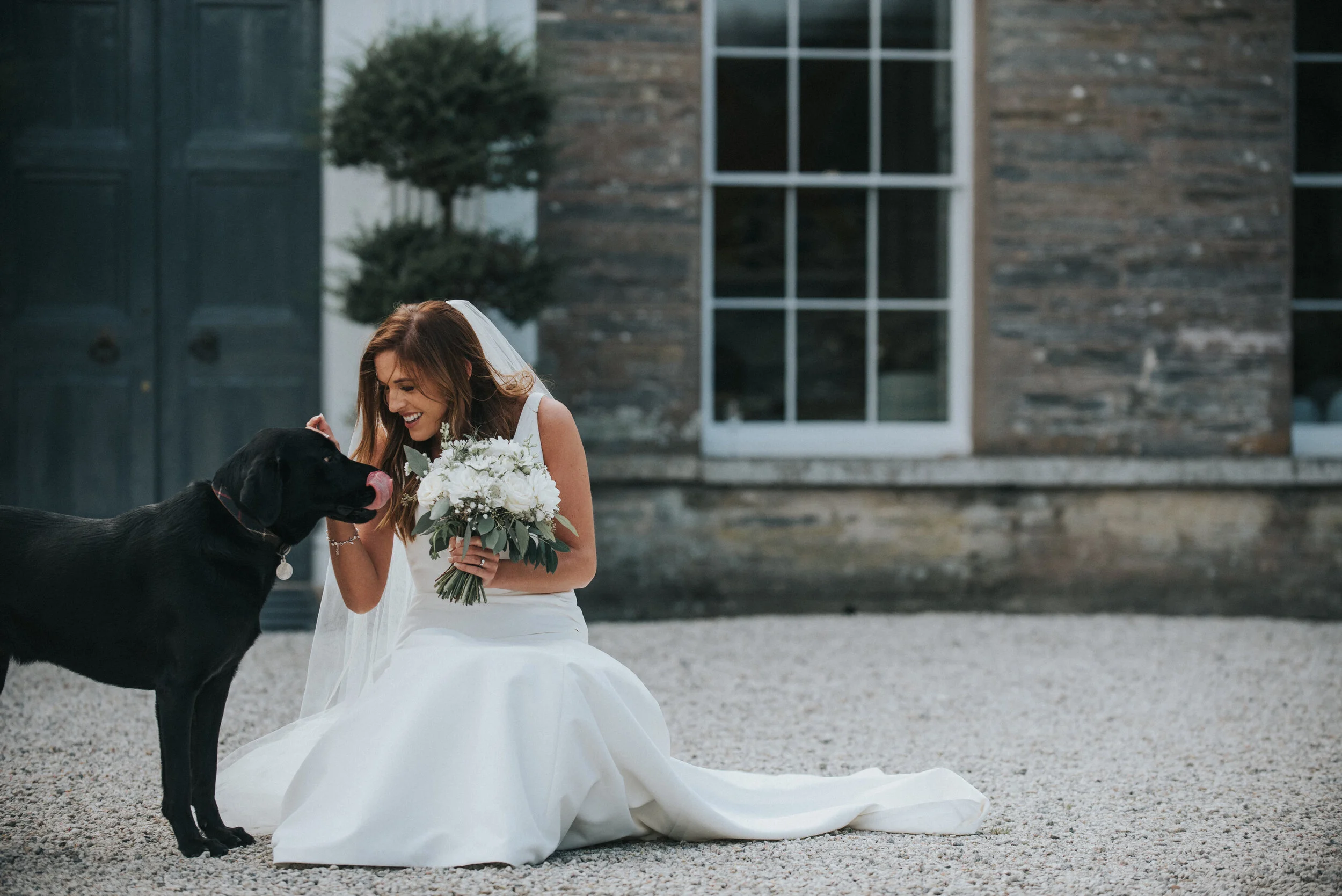 A bride in a white wedding dress kneels on gravel ground, smiling and petting her black dog while holding a bouquet of white flowers, outside a building with stone walls and large window.