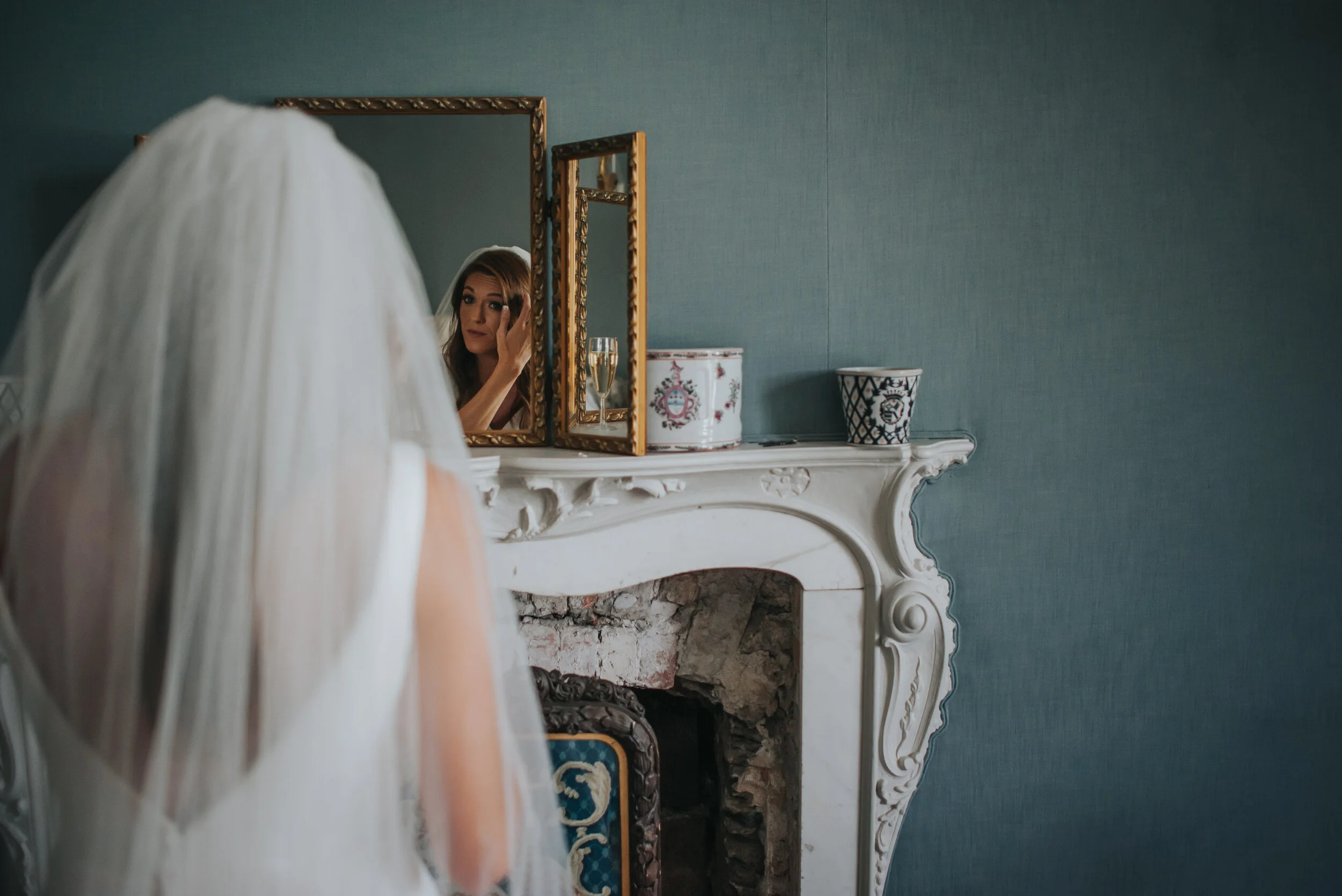 A woman wearing a veil is looking at herself in an ornate mirror as she adjusts her makeup, with a fireplace and decorative ceramics on the mantel behind her.