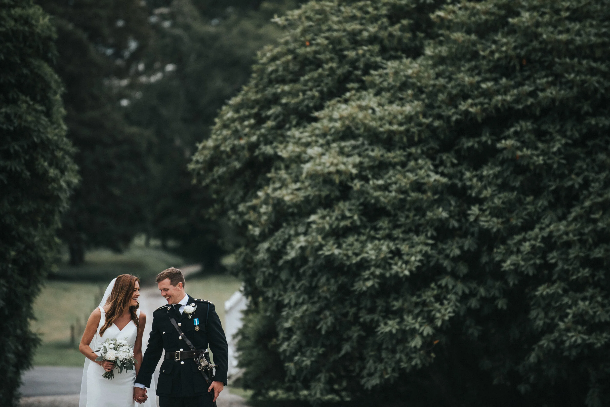 A bride and groom in wedding attire walking outdoors, holding hands and smiling, surrounded by lush greenery.
